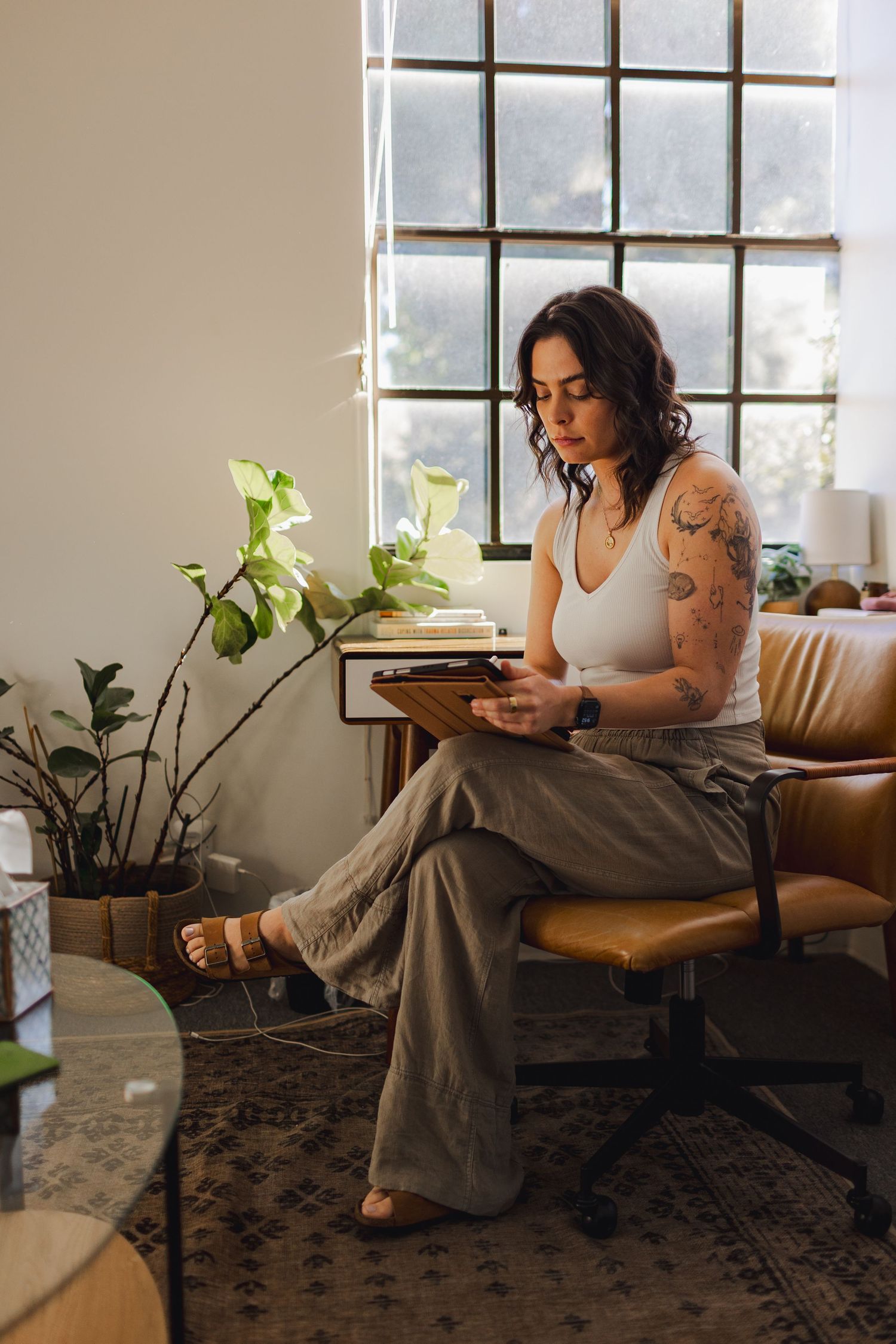 Tattooed woman in white tank top and olive pants reading a tablet in a cozy, plant-filled room by a large window.