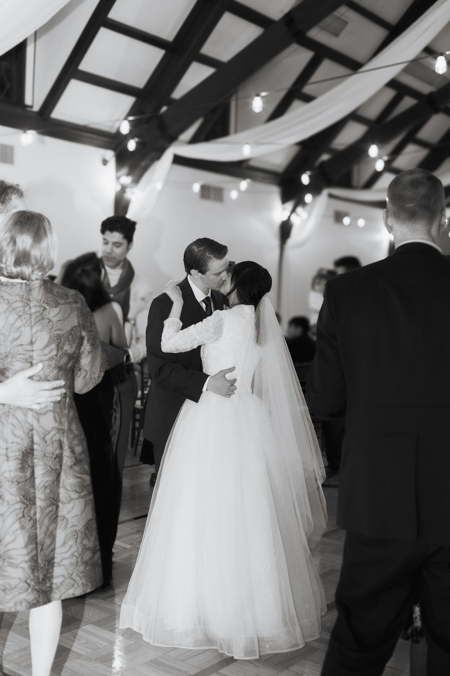 A black and white reception photo capturing a first dance under exposed wooden beams and string lights.