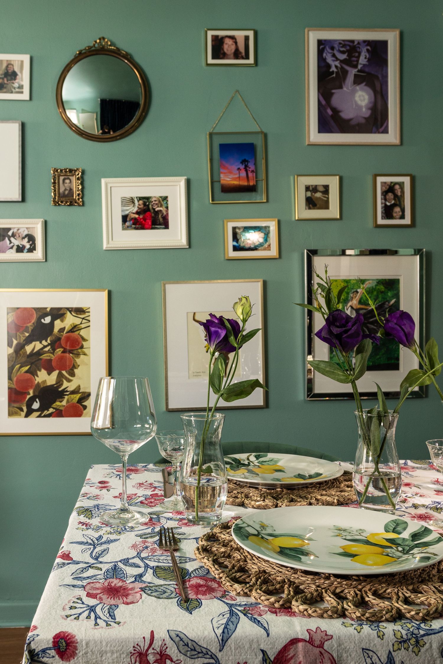 Eclectic dining room with green walls, gallery wall of framed art, floral tablecloth, and purple flowers in vases.