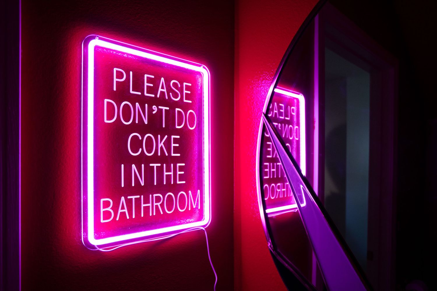 Pink neon sign reading 'Please Don't Do Coke In The Bathroom' mounted on a dark red wall.
