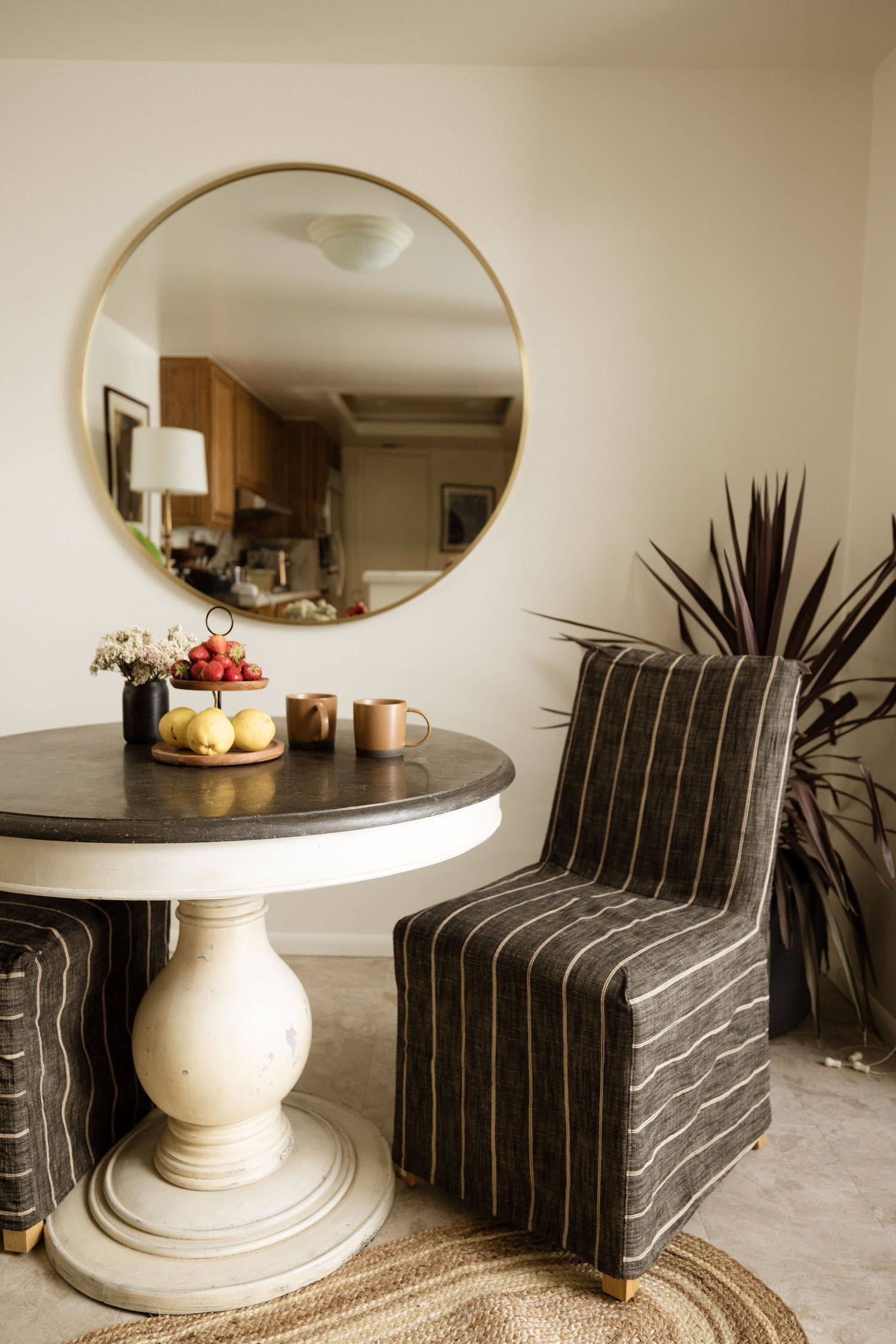 Round pedestal dining table with striped chairs, gold mirror, and fresh fruit centerpiece in a cozy dining nook.