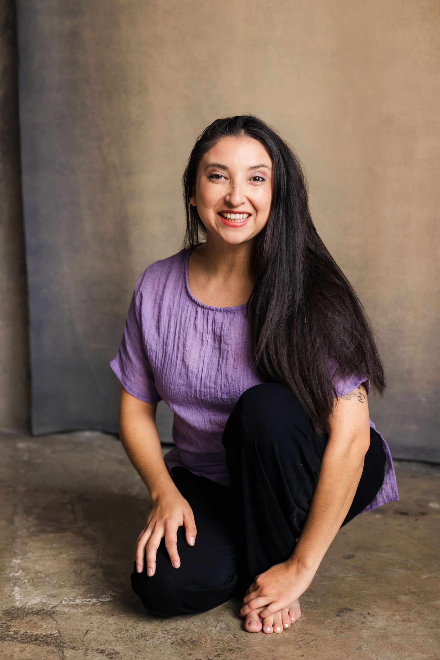 Smiling young woman with long dark hair sitting cross-legged on floor wearing purple top and black pants.