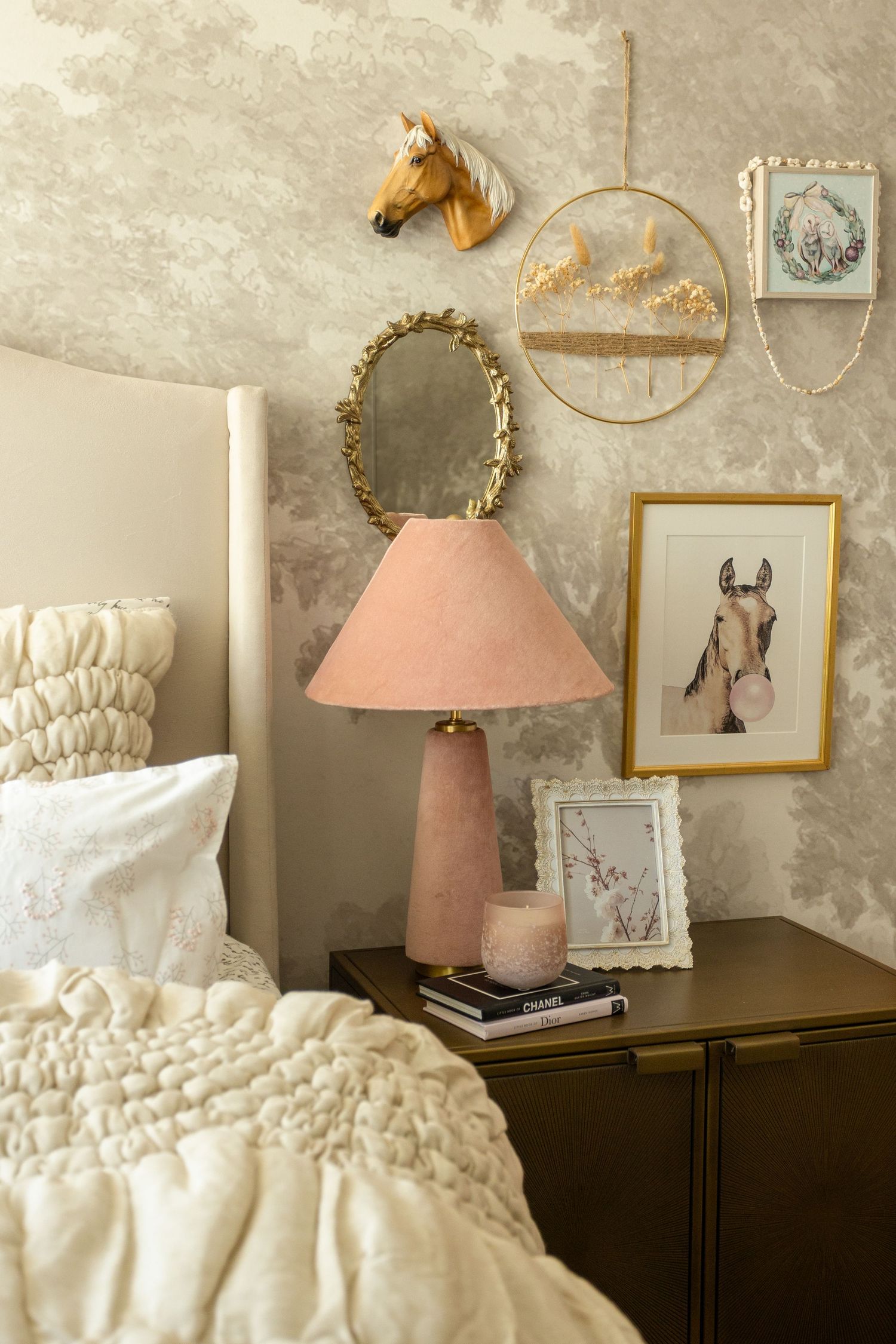 Pink lamp on dark nightstand beside bed with horse wall decor, gold mirror, and floral wallpaper in cozy bedroom.