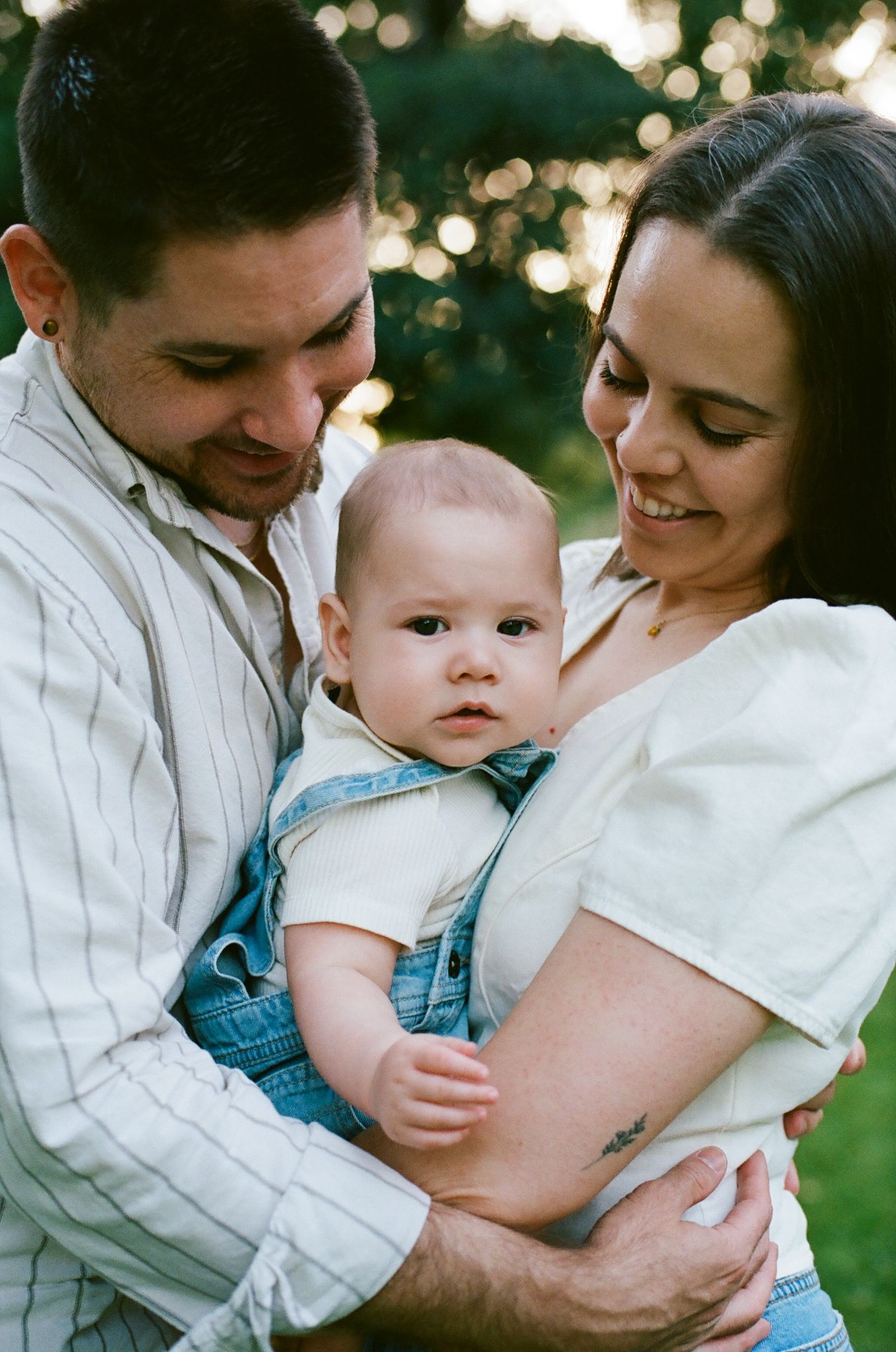 A young couple smiling and holding their baby in denim overalls outdoors in a warm, sunlit park setting.