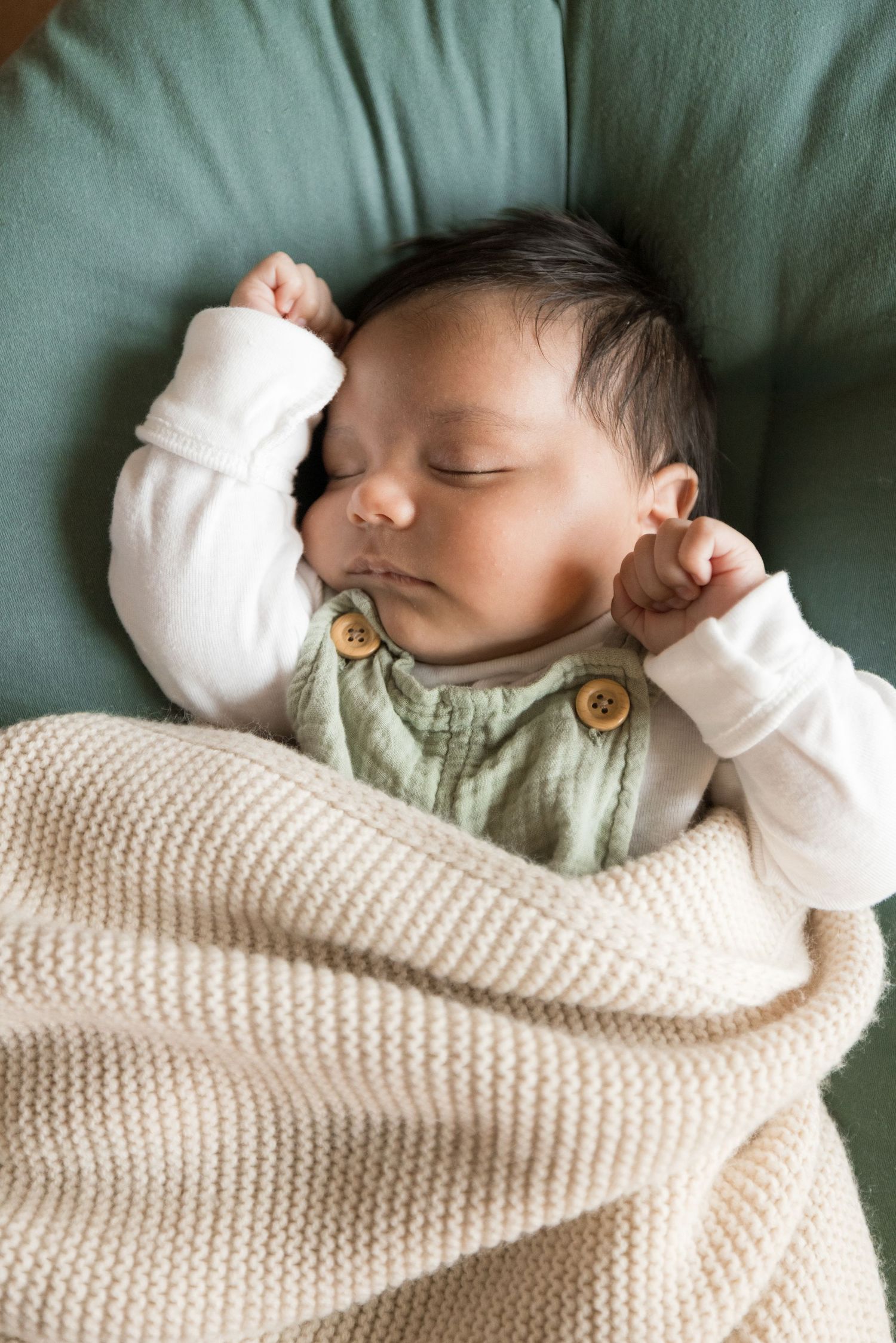 Sleeping newborn wrapped in cream knit blanket on green cushion.