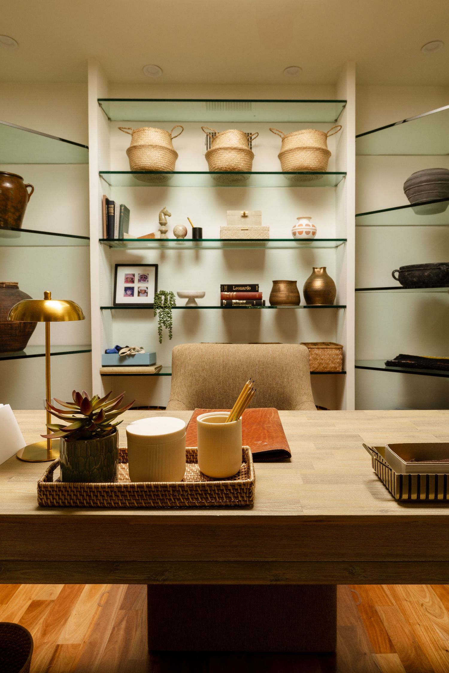 Elegant home office with glass shelves displaying woven baskets, vases, and decor, with a wooden desk featuring a succulent tray.