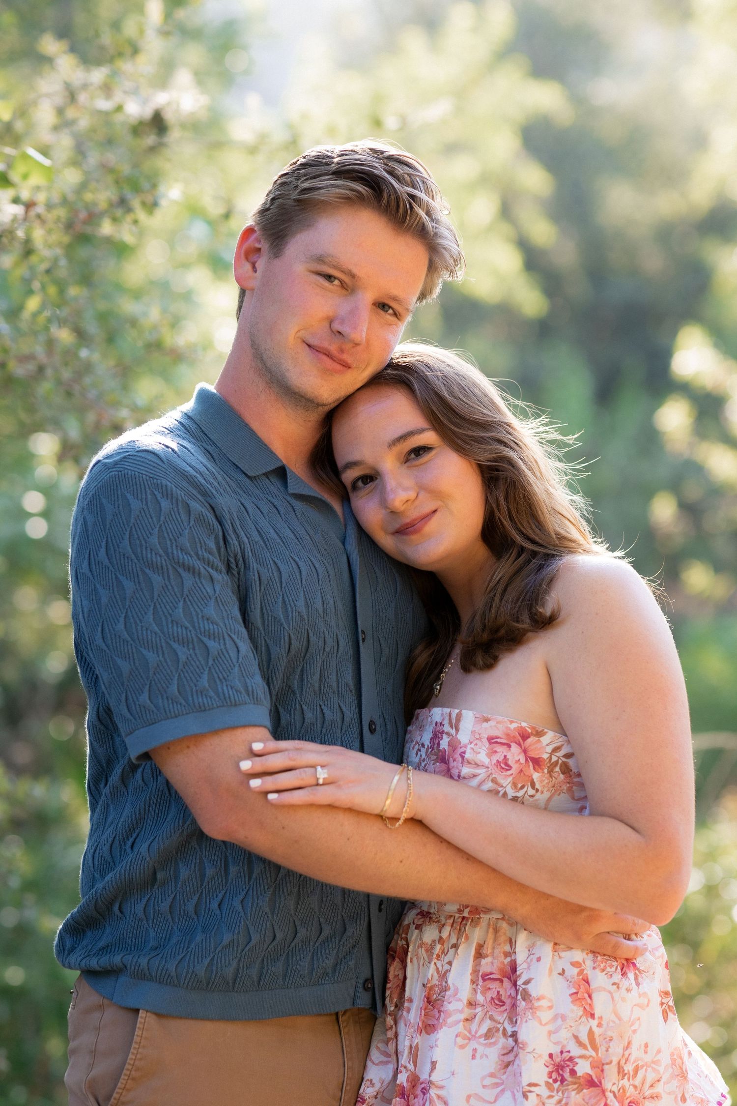 A young couple embraces outdoors, the woman in a floral dress leaning on the man in a blue knit shirt, with greenery behind them.