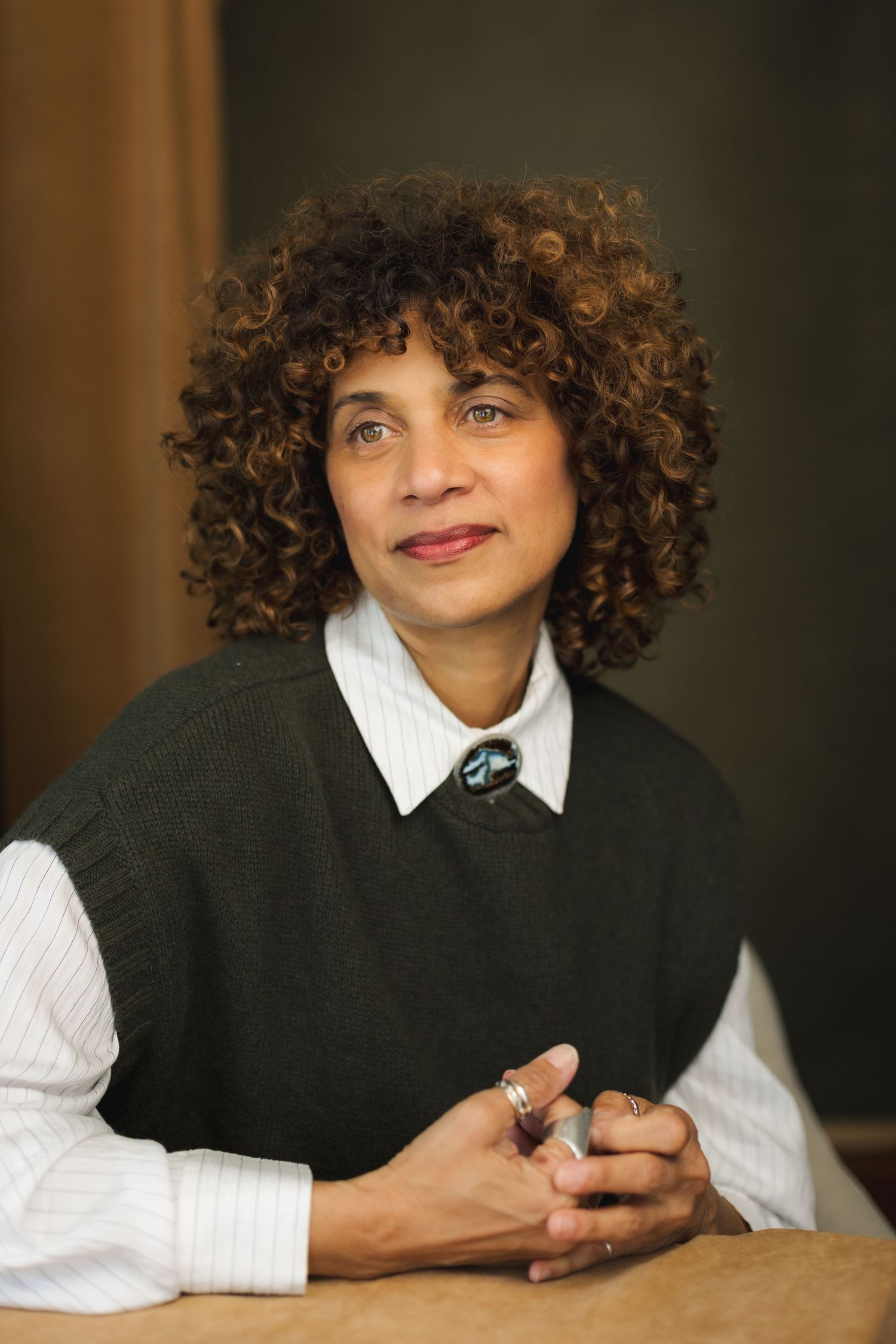 A woman with curly hair wearing a dark green vest and white shirt, sitting with hands clasped, smiling softly.