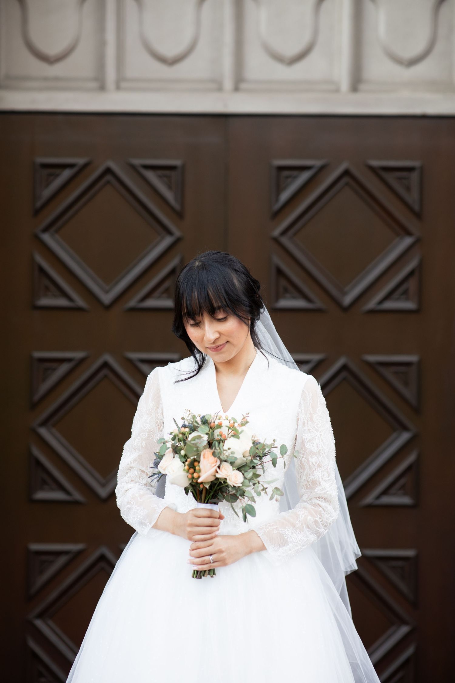 A bride in a white wedding dress holds a delicate bouquet against a dark wooden door background.