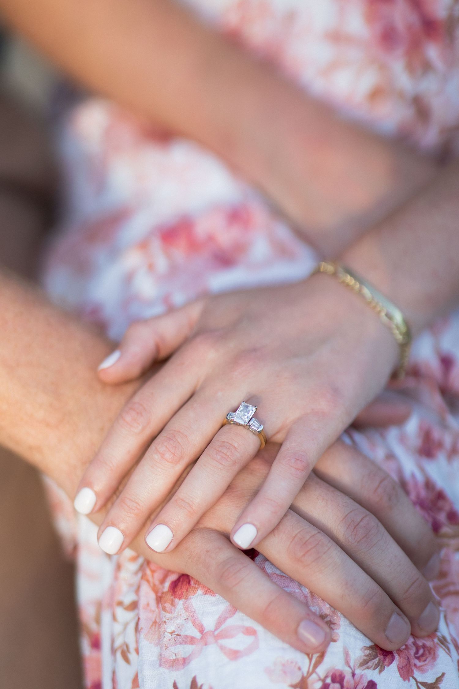 Close-up of hands showing an emerald-cut diamond engagement ring on ring finger, with pink floral dress background.