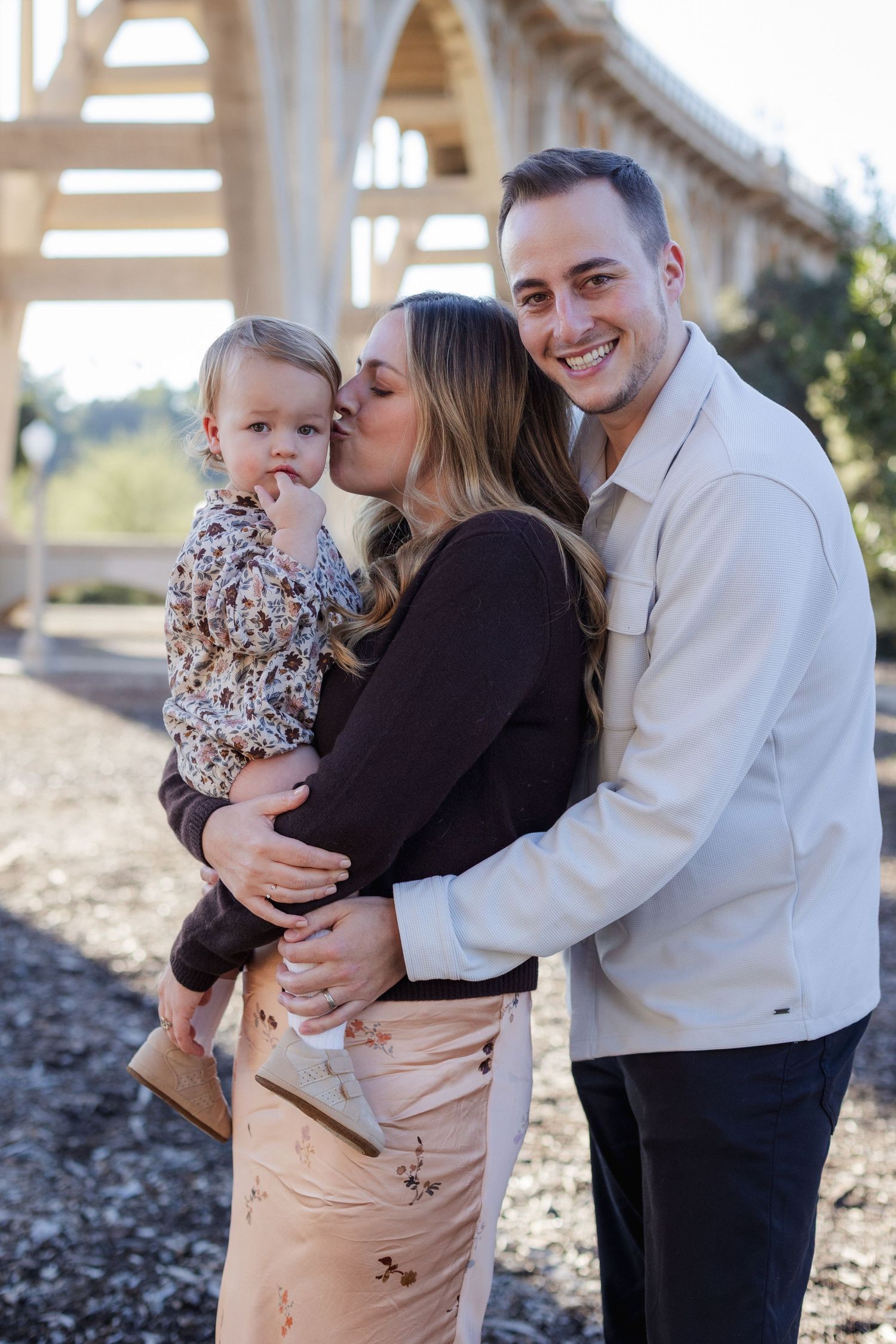 A smiling family poses outdoors near a bridge, with a woman kissing a toddler while a man embraces them both.