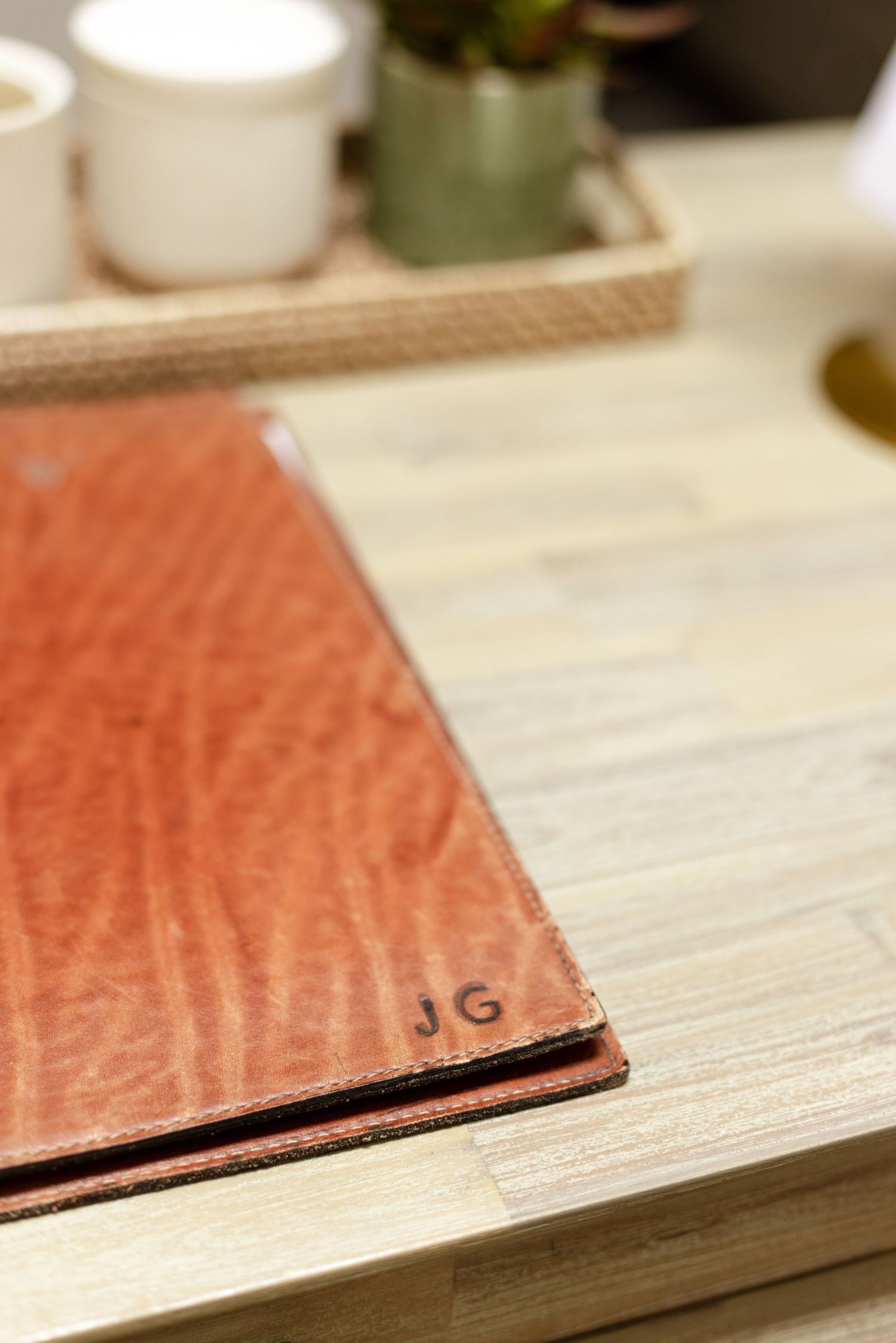 Close-up of a monogrammed brown leather portfolio with initials JG on a light wood desk surface.