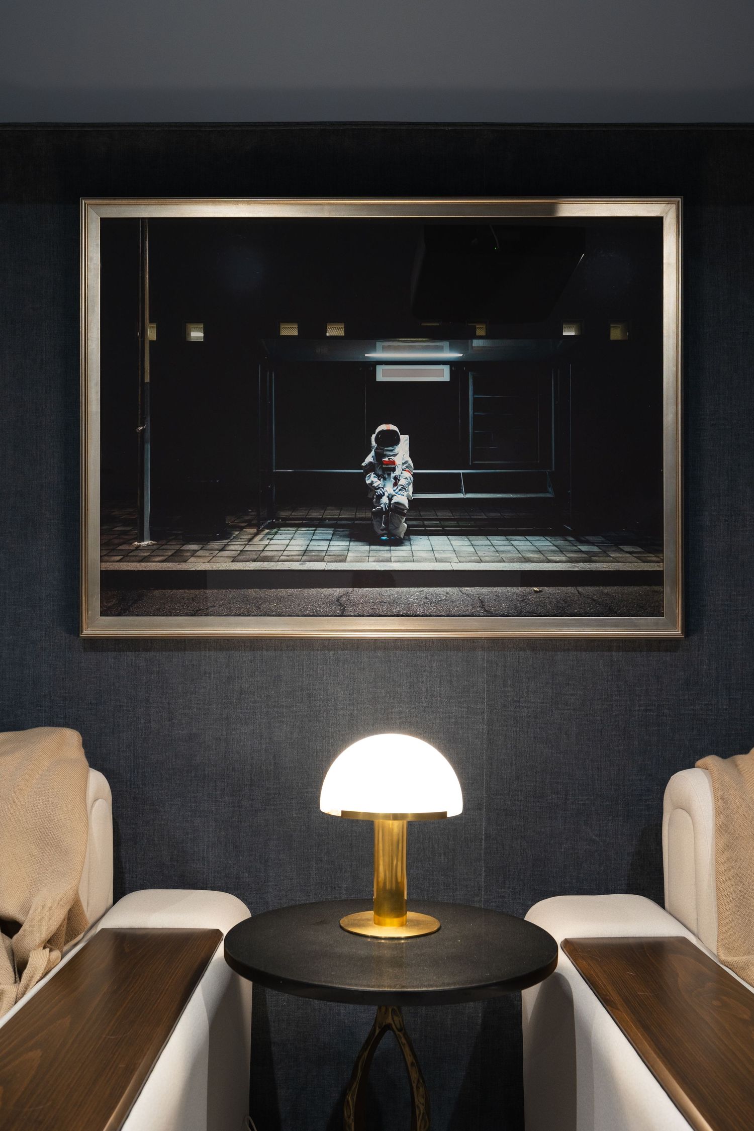 Framed astronaut photo above a glowing mushroom table lamp between two chairs in a dark, sophisticated lounge.