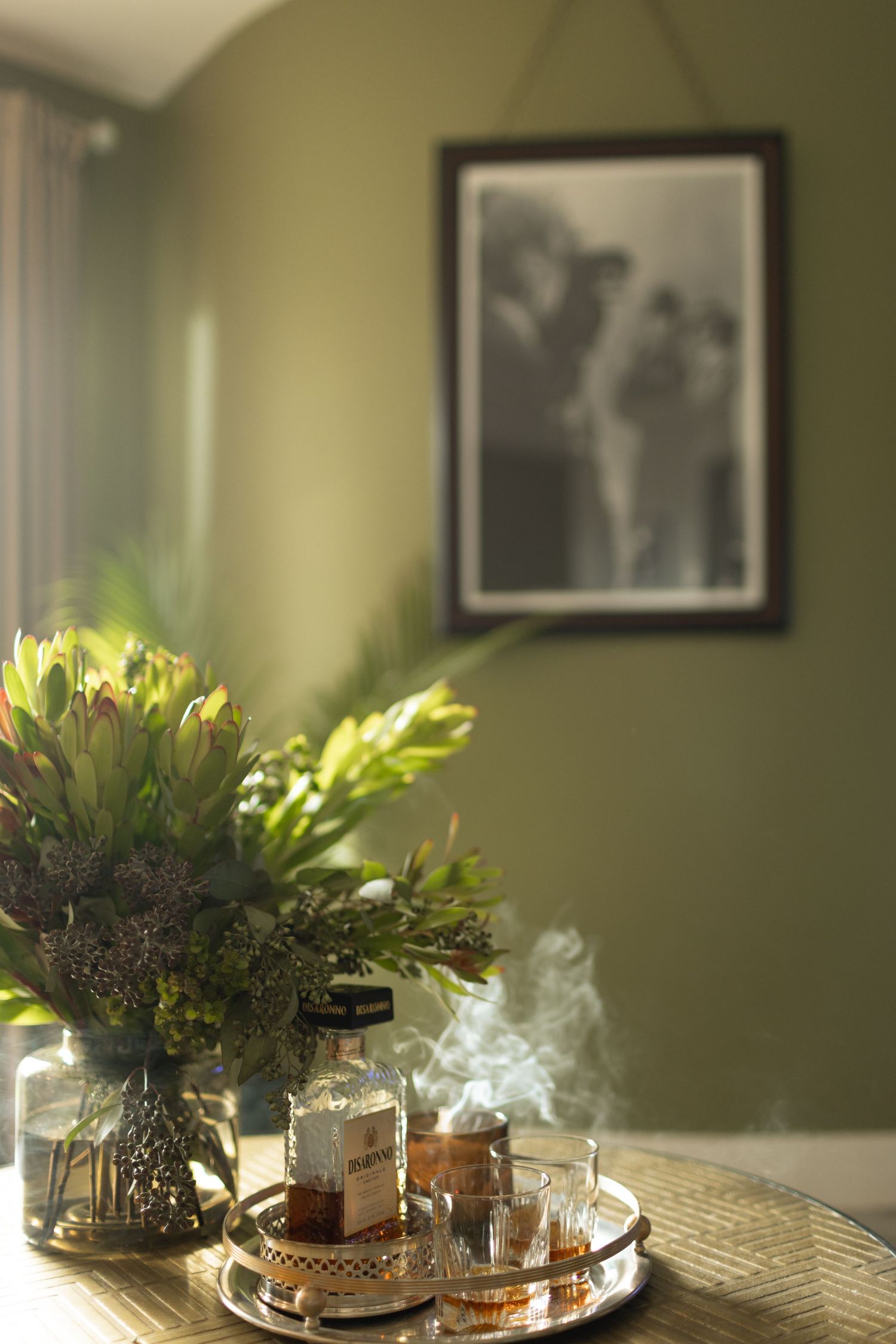 Golden tray with perfume bottles and smoking incense beside tropical flowers on table, green wall with black and white photo.