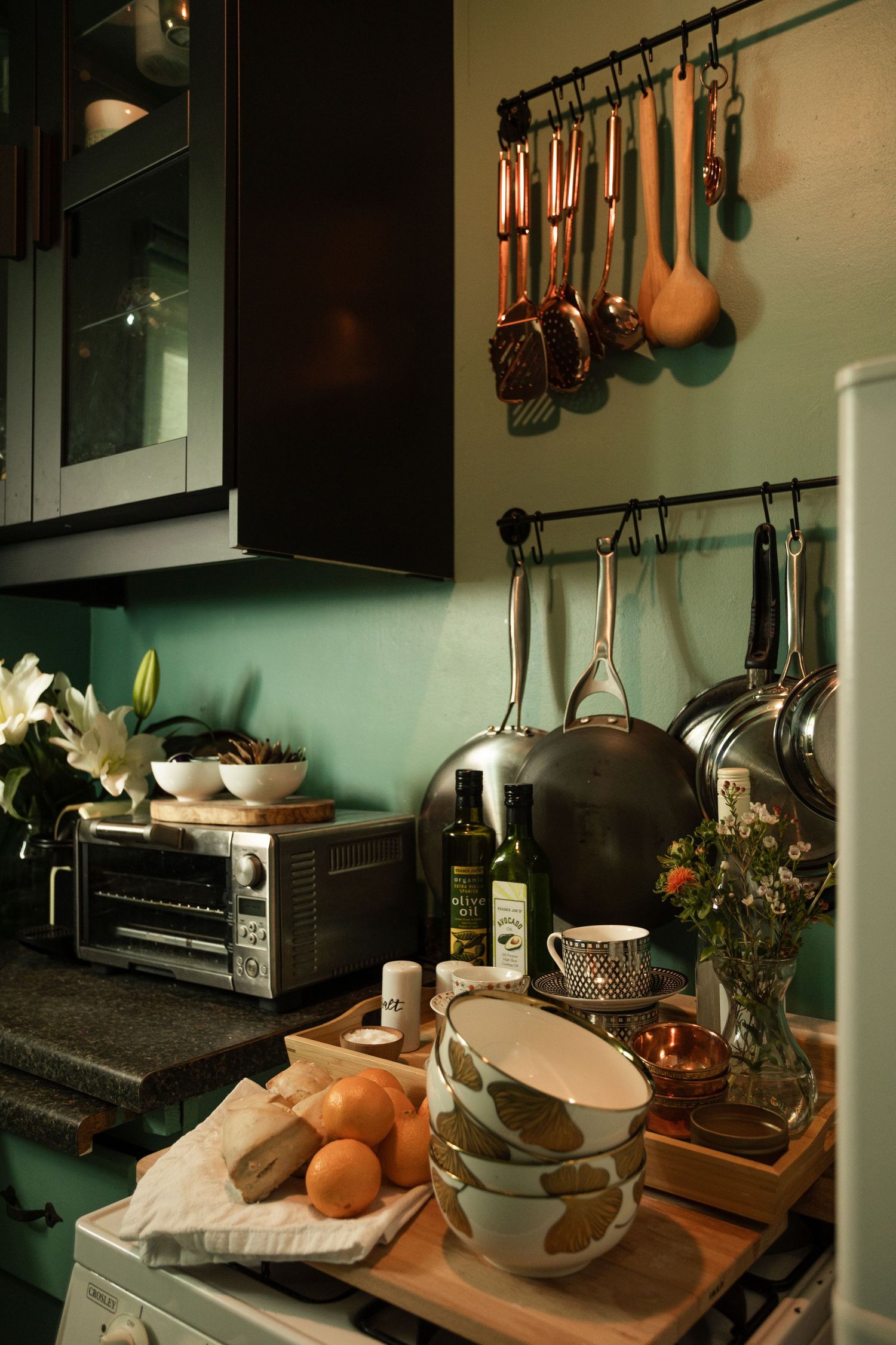 Cozy kitchen counter with copper utensils hanging on wall rack, toaster oven, olive oil, bowls, and fresh oranges on cutting board.