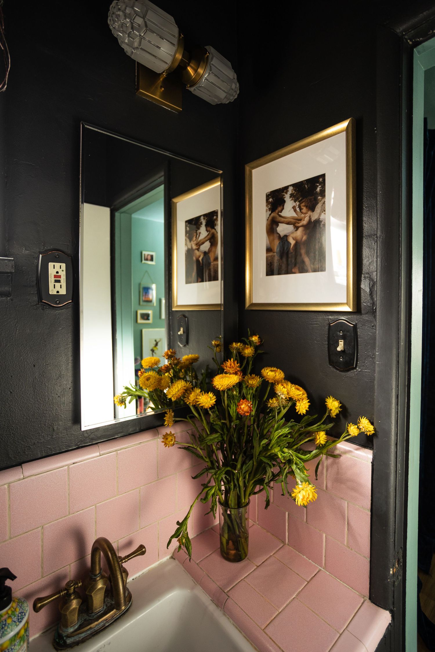 Dark moody bathroom with pink tile sink, yellow flowers, gold-framed art, and black walls.