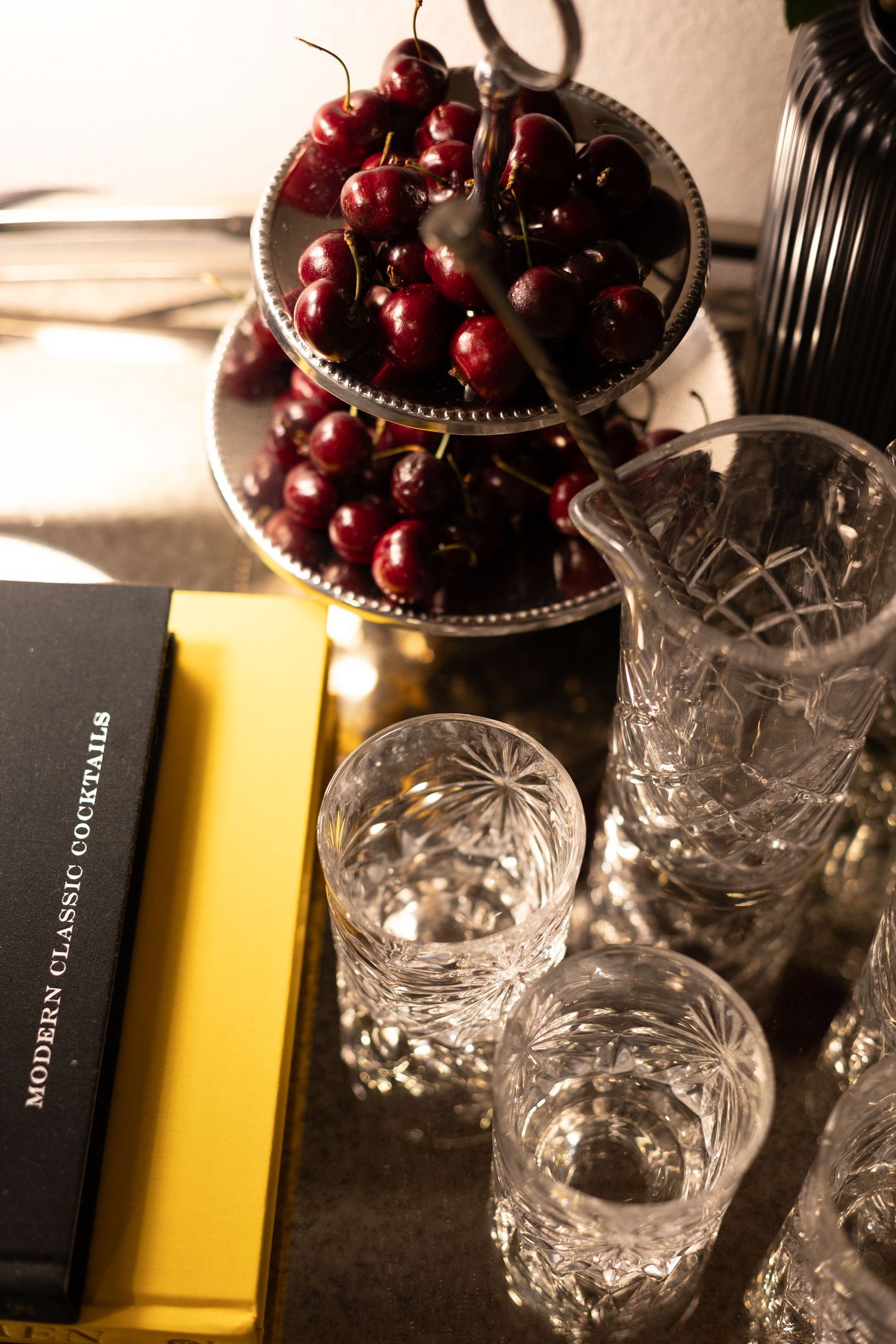 Crystal glassware and a pitcher beside fresh cherries on a tiered stand, with a Modern Classic Cocktails book on a golden tray.