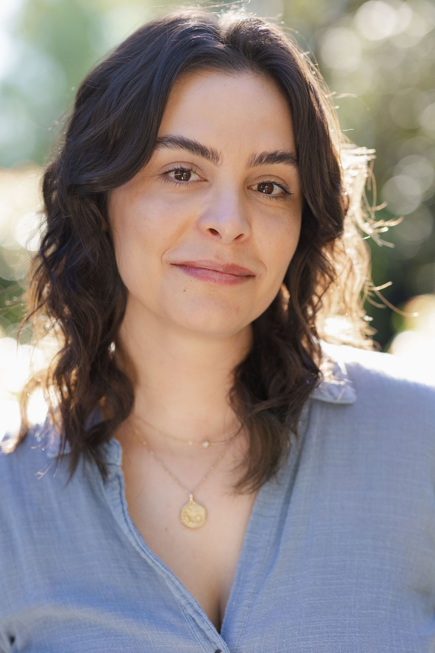 Portrait of a young woman with dark wavy hair wearing a blue shirt and gold pendant necklace outdoors.