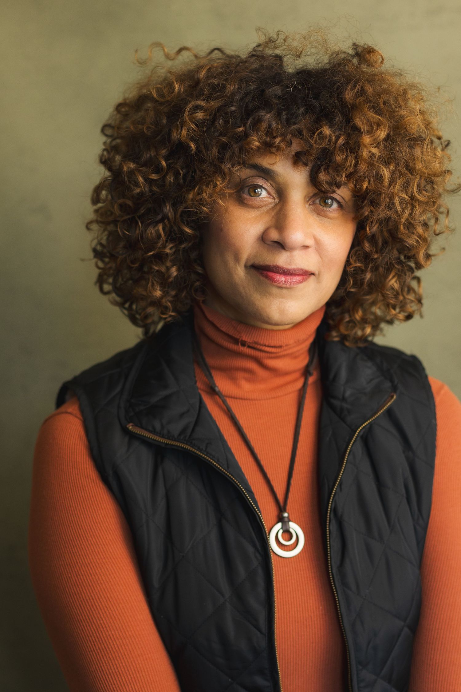 Woman with curly auburn hair wearing an orange turtleneck, black vest, and circular pendant necklace.