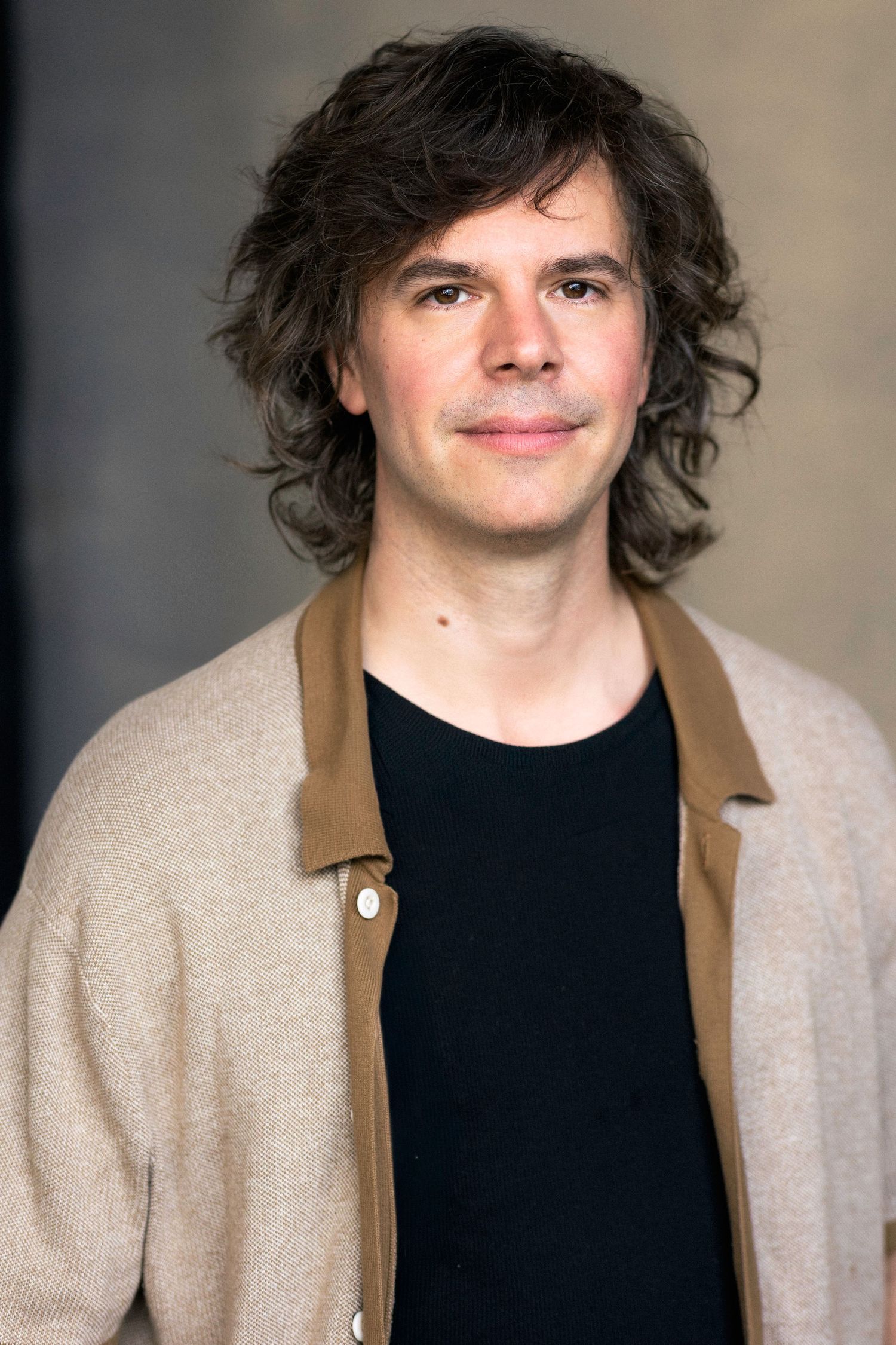 Smiling man with curly dark hair wearing a black shirt and beige cardigan poses for a professional headshot.