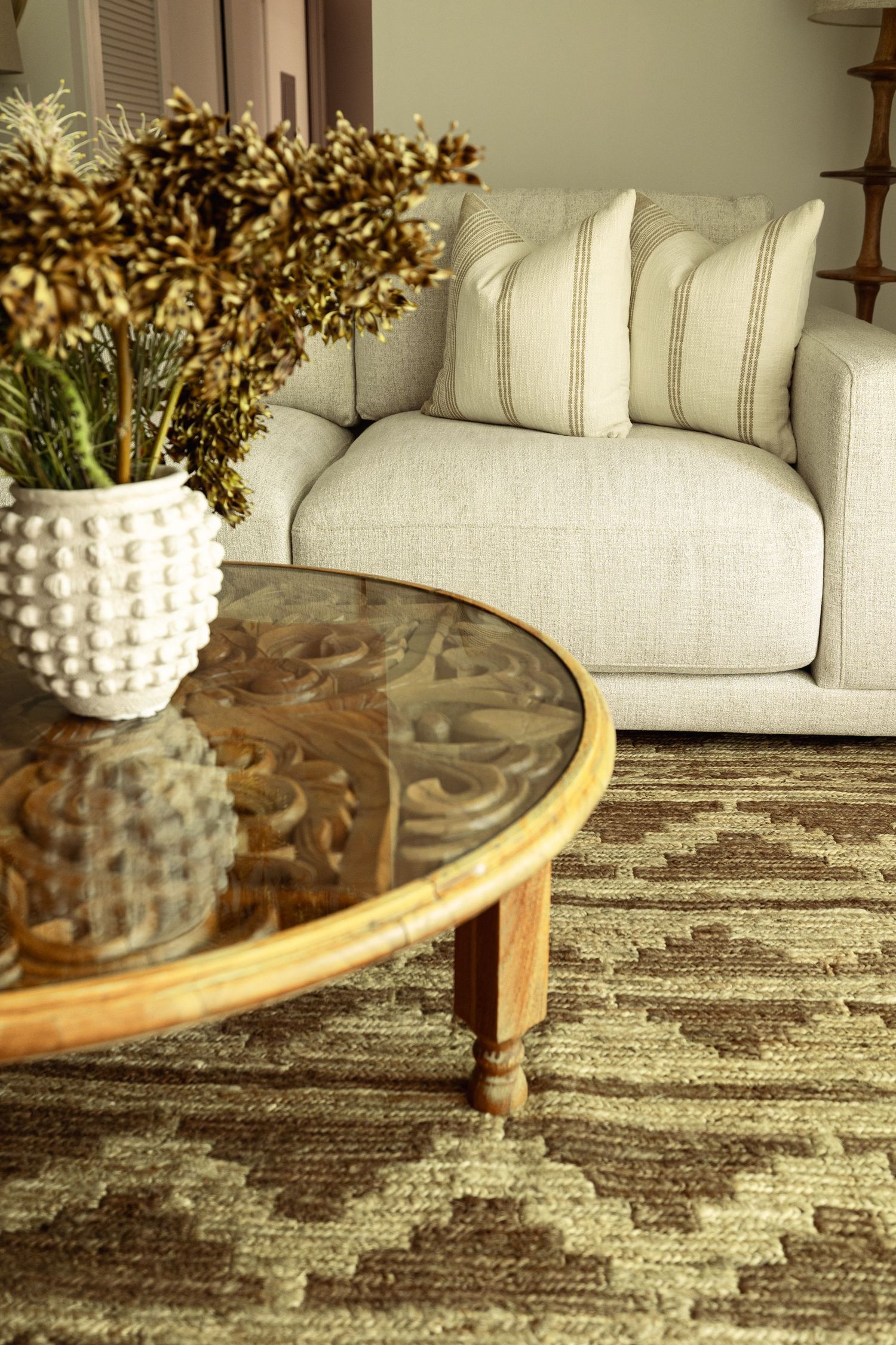 Cozy living room with beige linen sofa, striped pillow, round glass coffee table, white textured vase, and brown geometric rug.