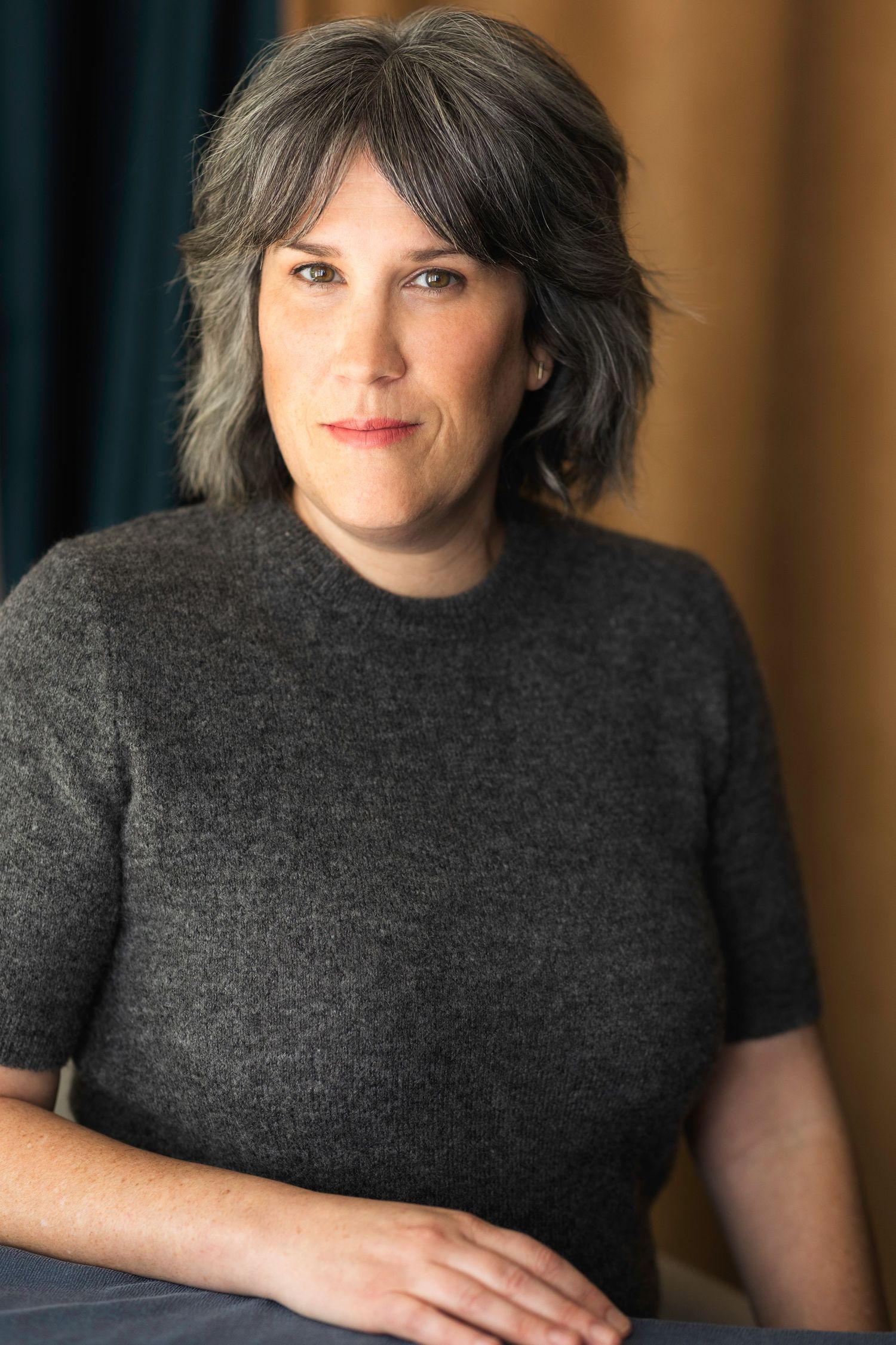 Woman with gray-streaked hair wearing a dark gray sweater, seated and looking at camera in a warm indoor setting.