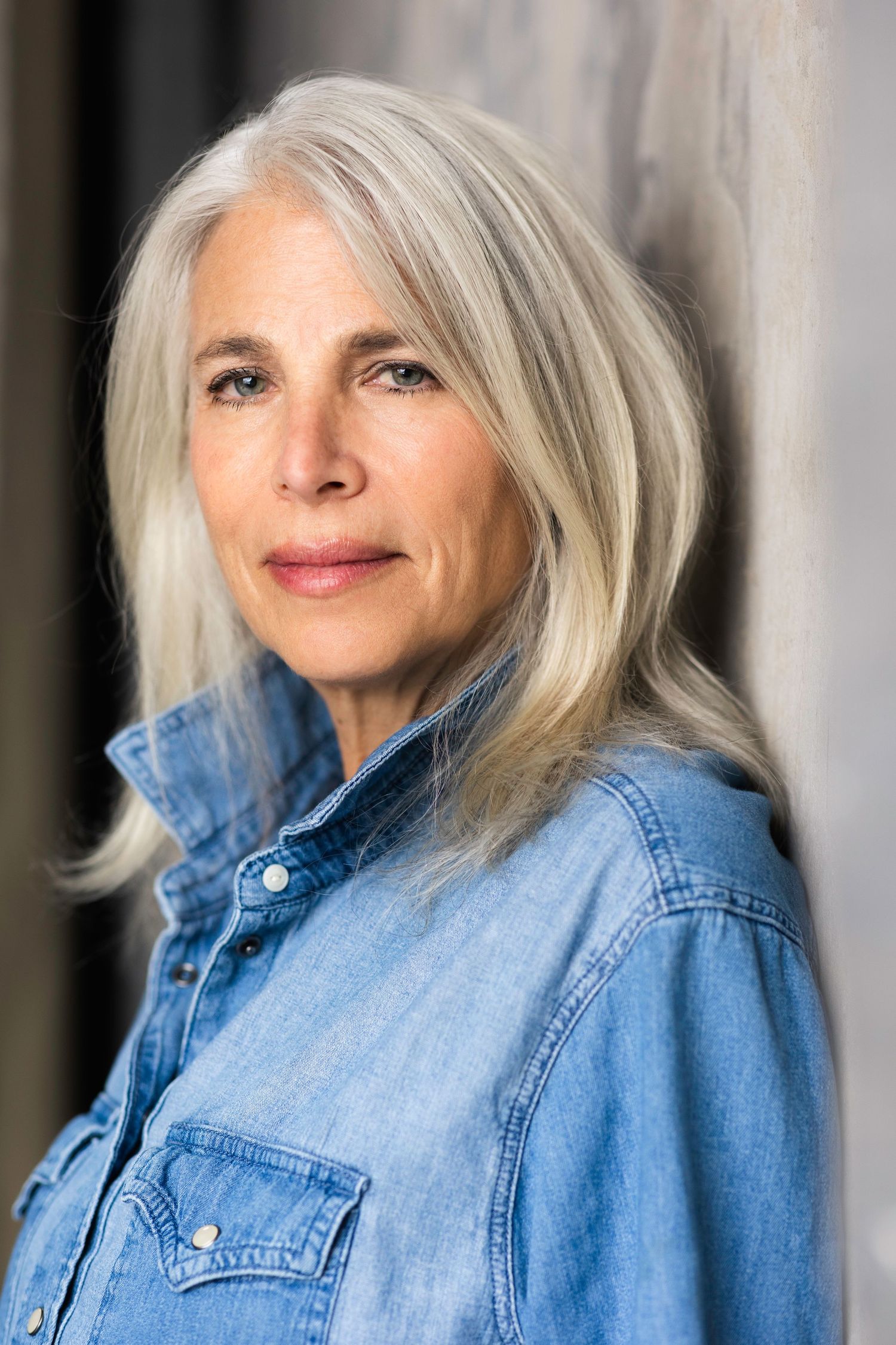 Mature woman with silver hair wearing a denim shirt, posing confidently against a gray wall.