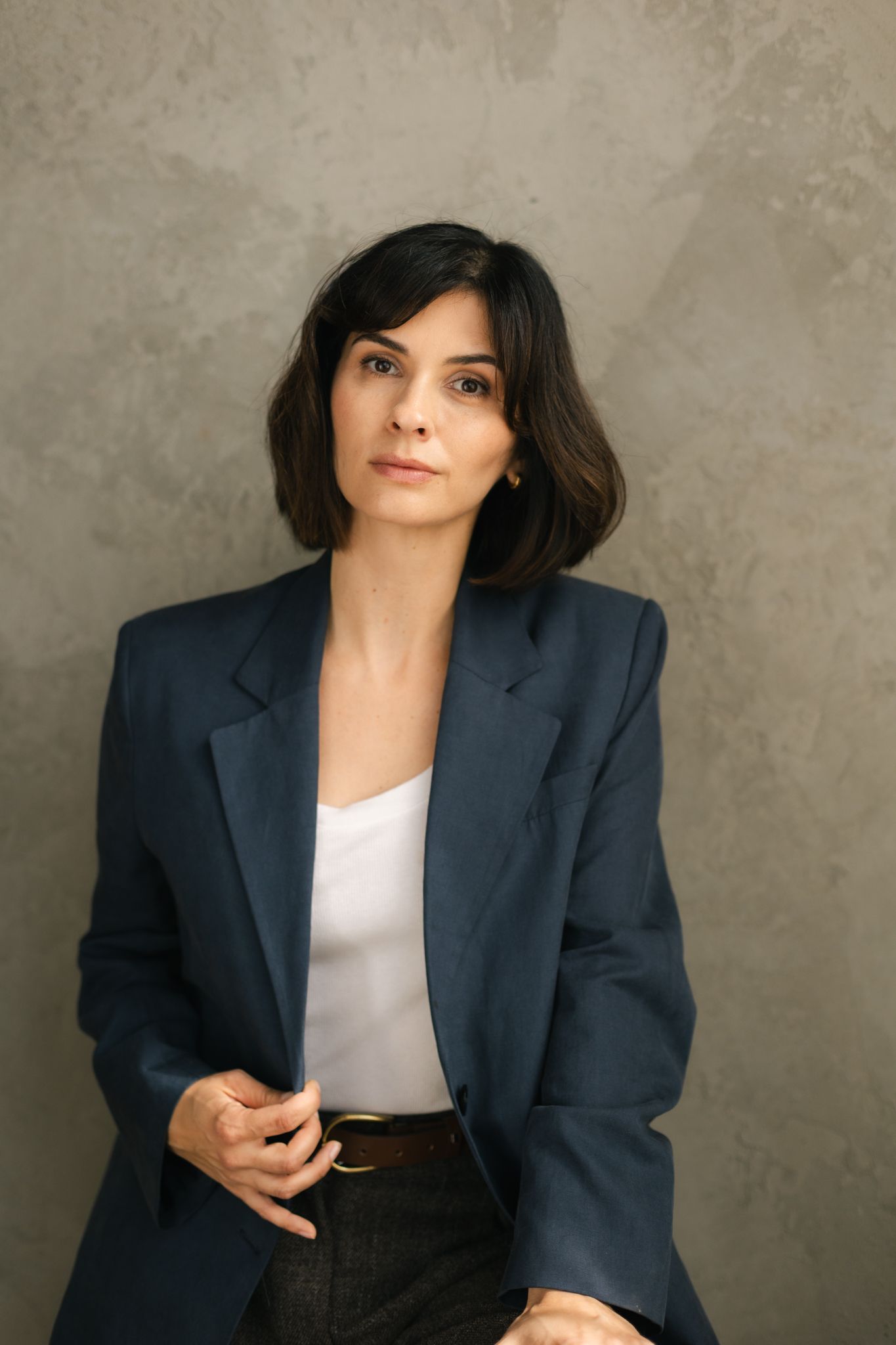 Woman in navy blazer and white top posing against textured grey wall.