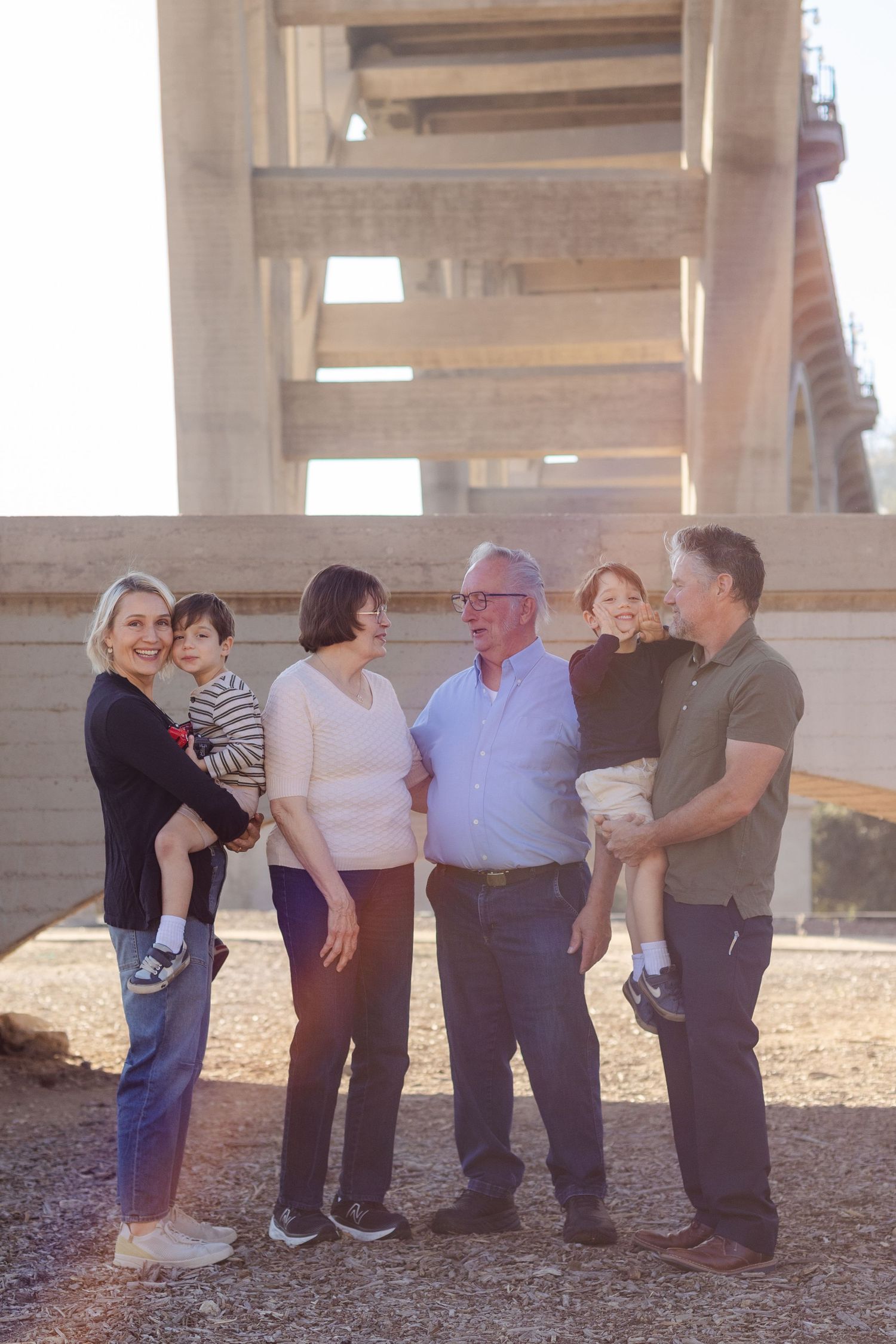 Multi-generational family of six smiling together outdoors beneath a large concrete bridge structure.