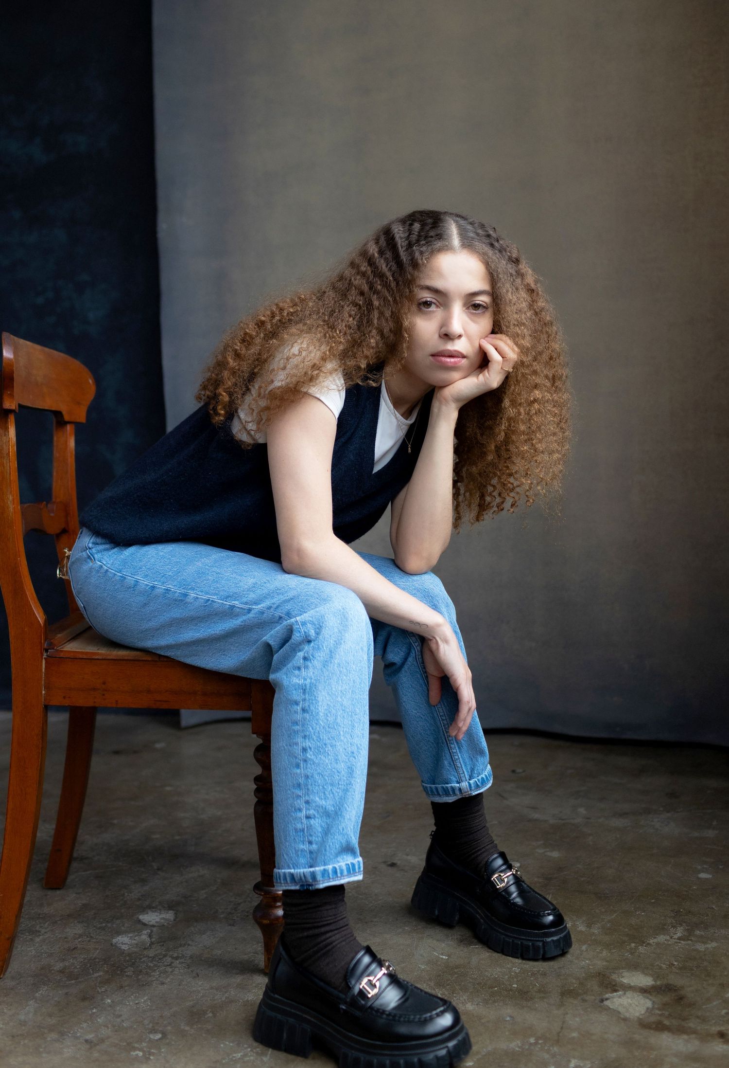 Person in black vest and jeans sits casually on wooden chair.