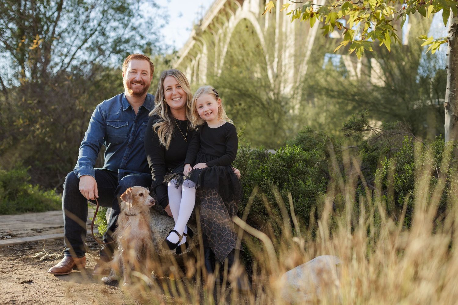 Family of three with a golden dog posing outdoors near an arched bridge surrounded by nature.