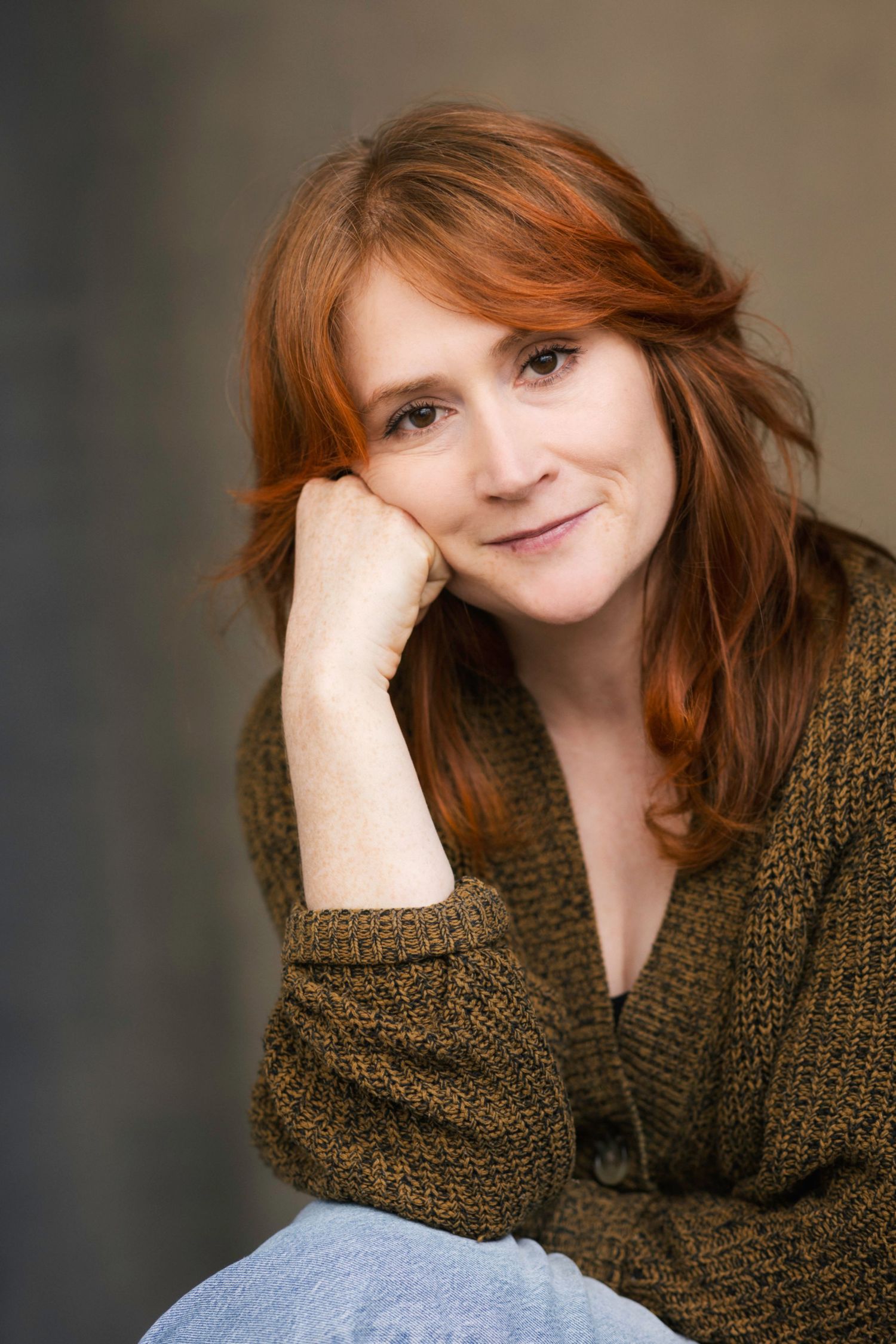 A smiling redhead woman in a brown knit cardigan rests her chin on her hand in a casual portrait photo.