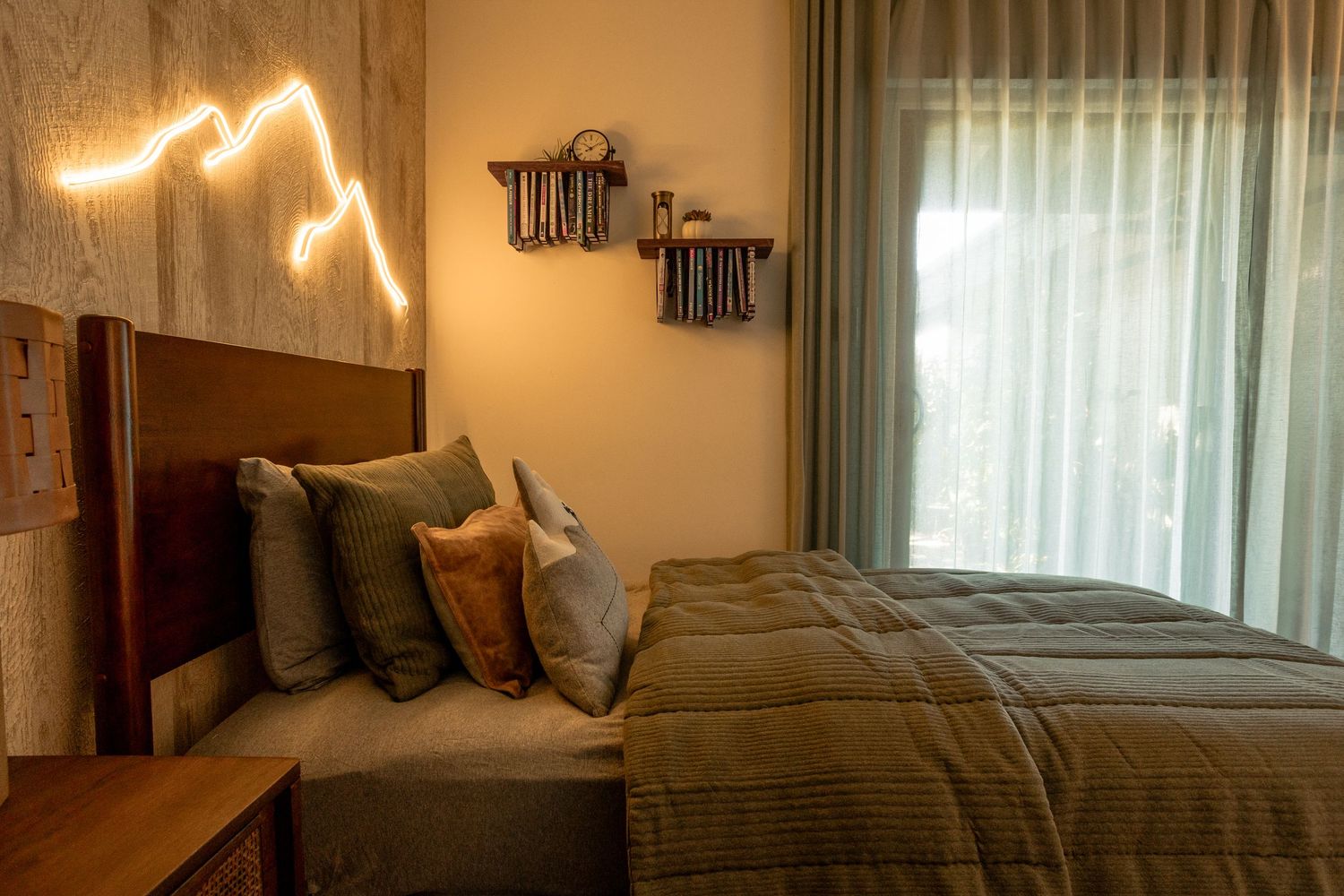 Cozy bedroom with green bedding, decorative pillows, mountain neon light art, wooden headboard, and sheer curtains.