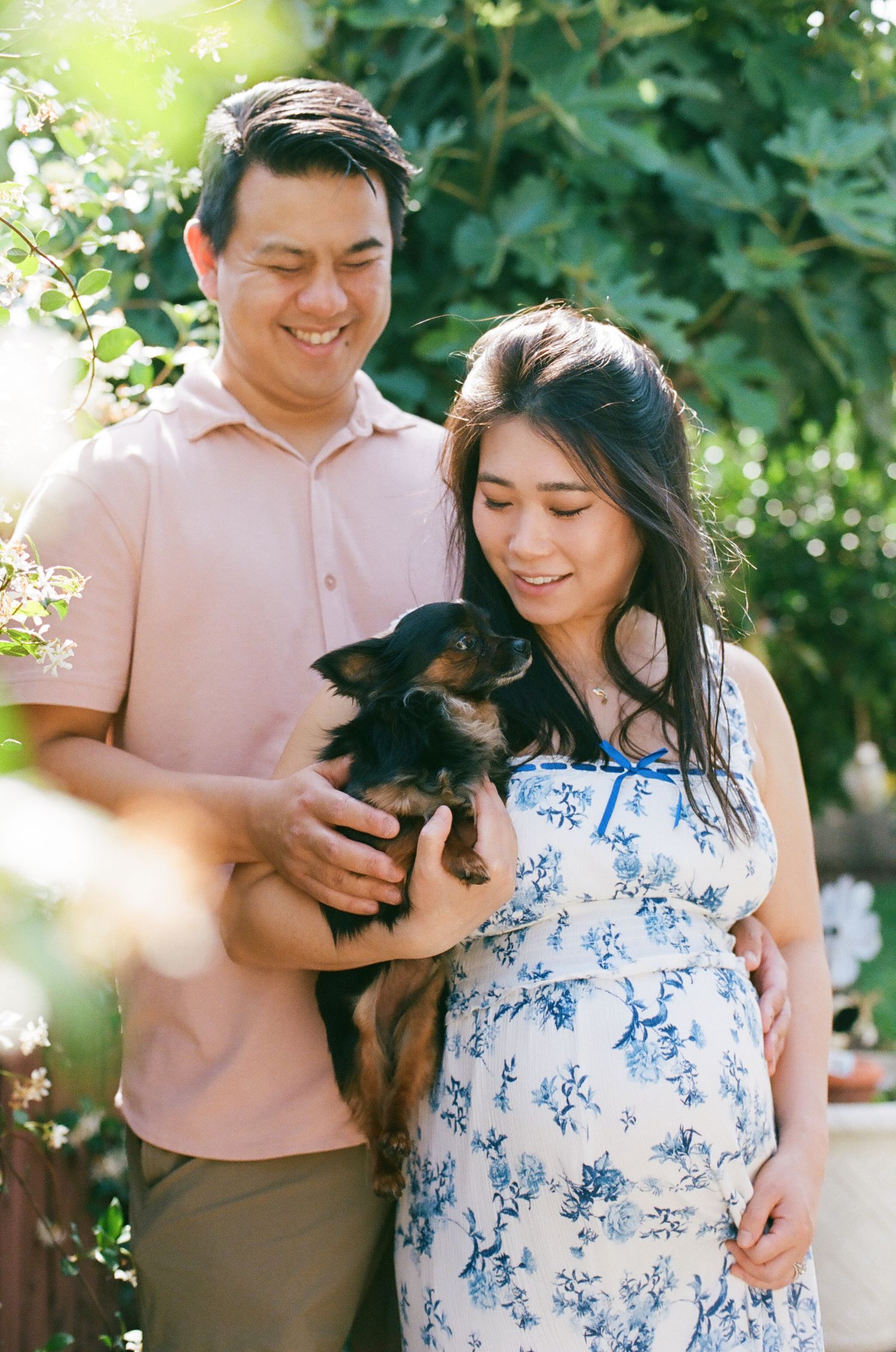 Smiling couple holding a small black dog outdoors, woman in floral dress with baby bump, lush green garden background.
