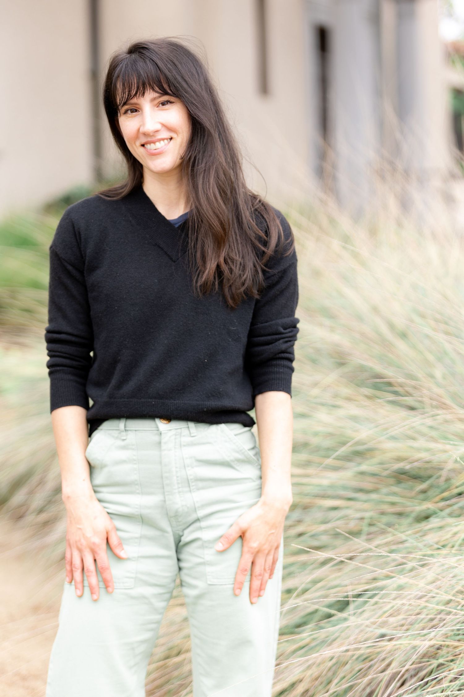 A smiling woman with long dark hair wearing a black sweater and light green pants standing outdoors near tall grass.