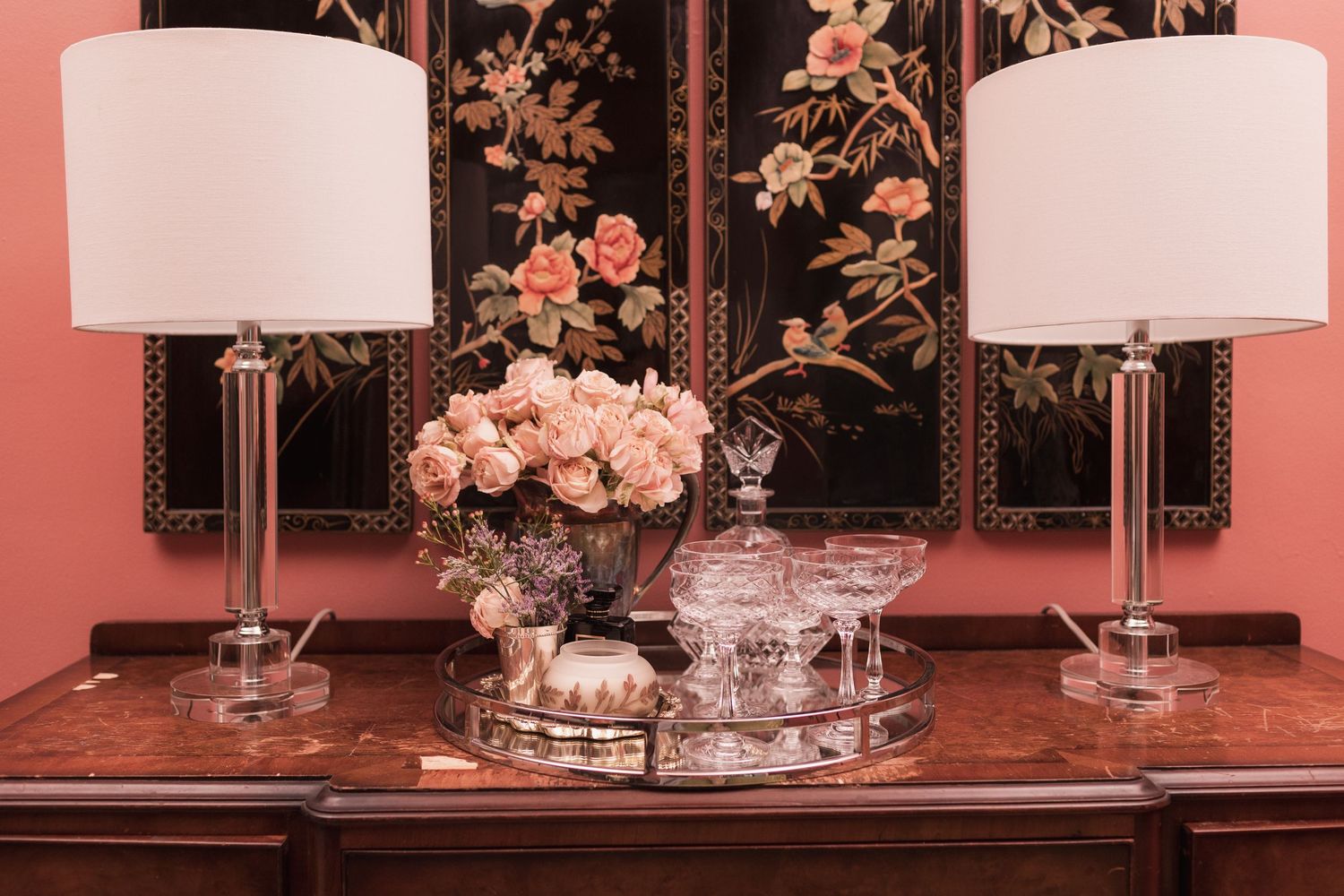 Elegant sideboard with crystal lamps, pink roses, and glassware on a mirrored tray against a pink wall with floral screen.