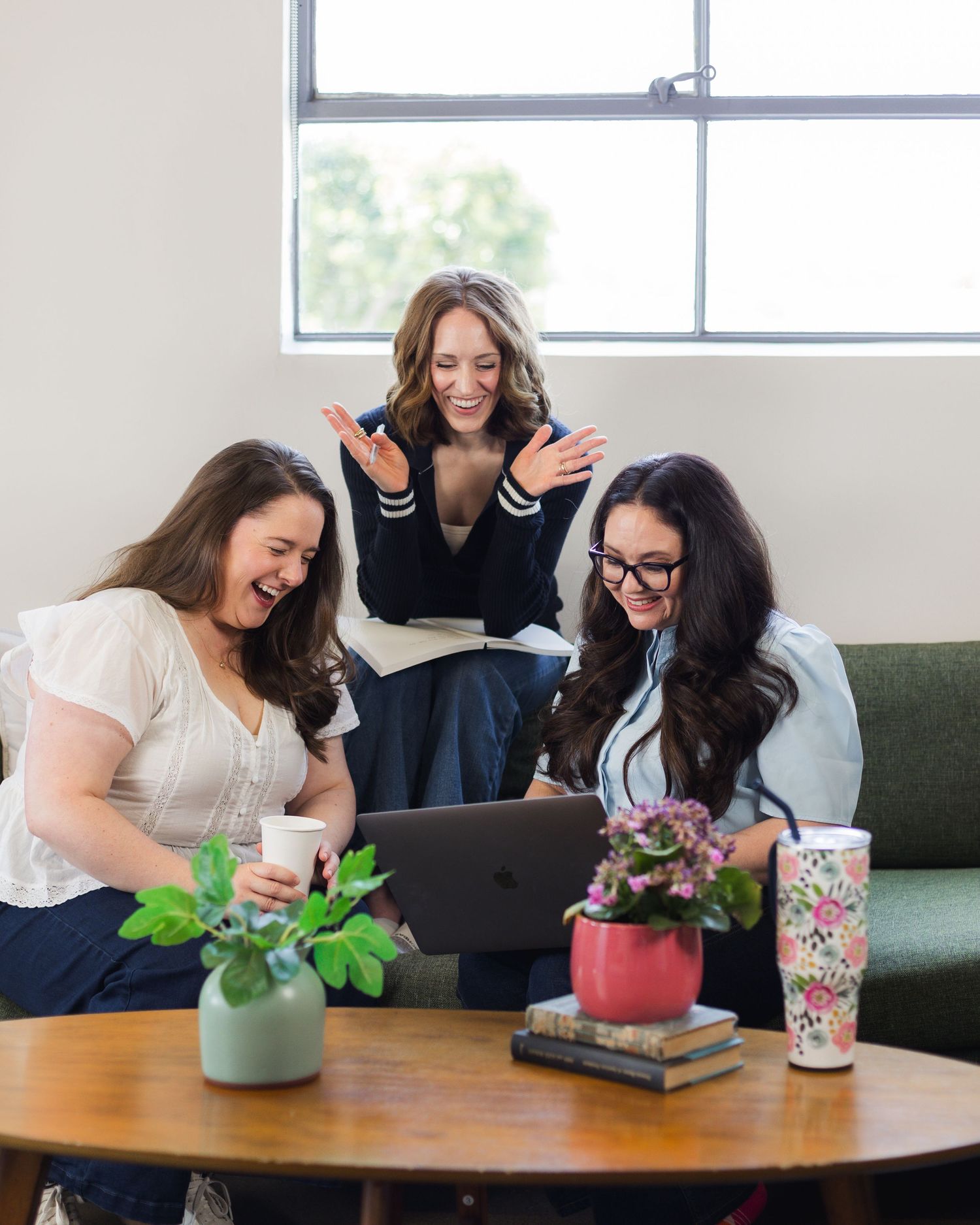 Three women laugh and react excitedly while looking at a laptop on a wooden coffee table in a bright, modern office.