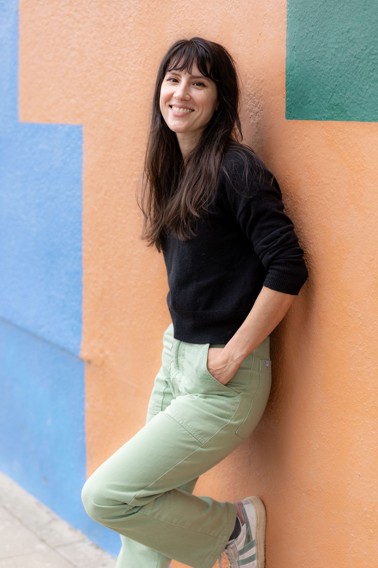 A smiling woman with dark hair leans against a colorful orange wall, wearing a black sweater and light green pants.