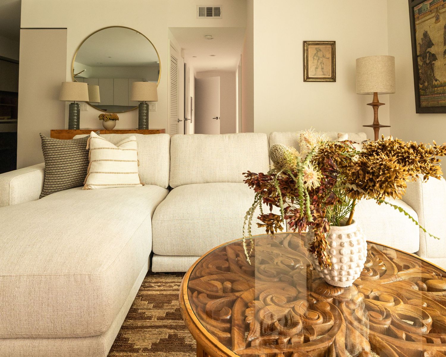 Cream sectional sofa with patterned rug and ornate wooden coffee table with dried floral arrangement.