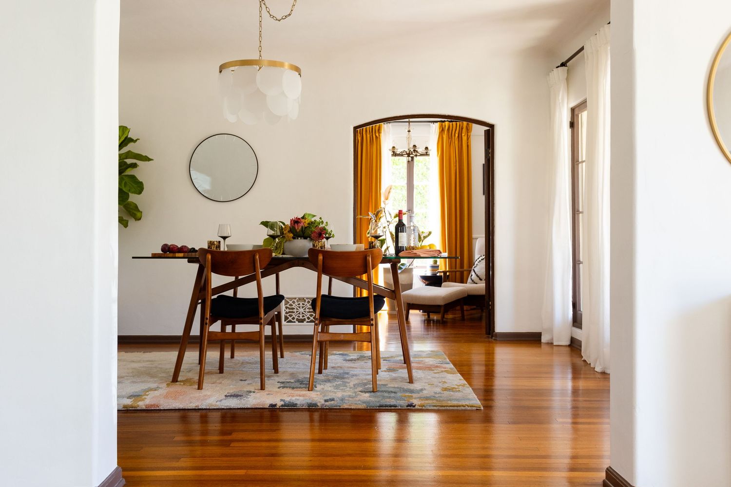 Mid-century modern dining room with hardwood floors, wooden chairs, floral rug, arched window, and warm amber curtains.