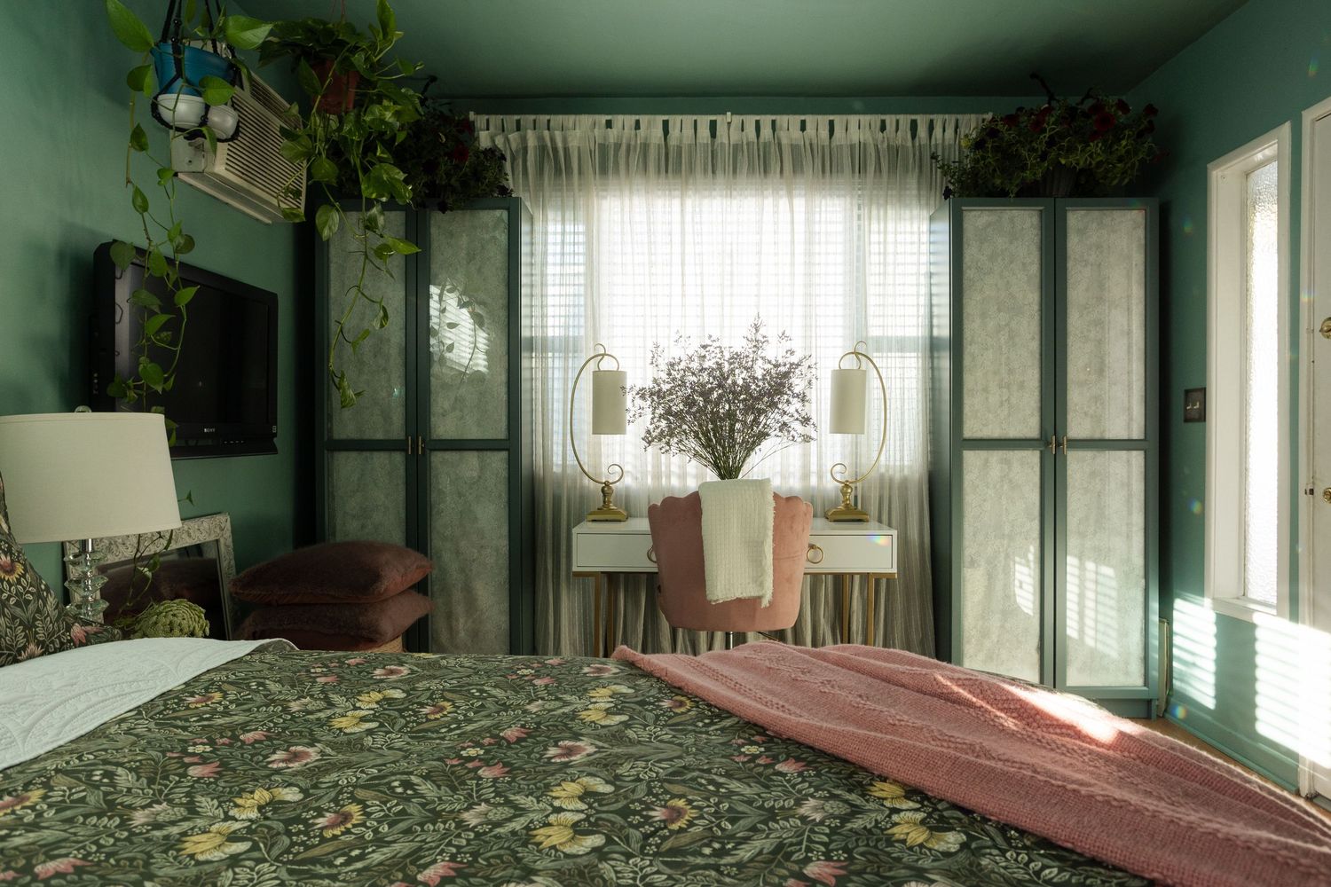 Green bedroom with floral bedspread, built-in wardrobes, vanity desk, sheer curtains, and lush plants near a sunlit window.