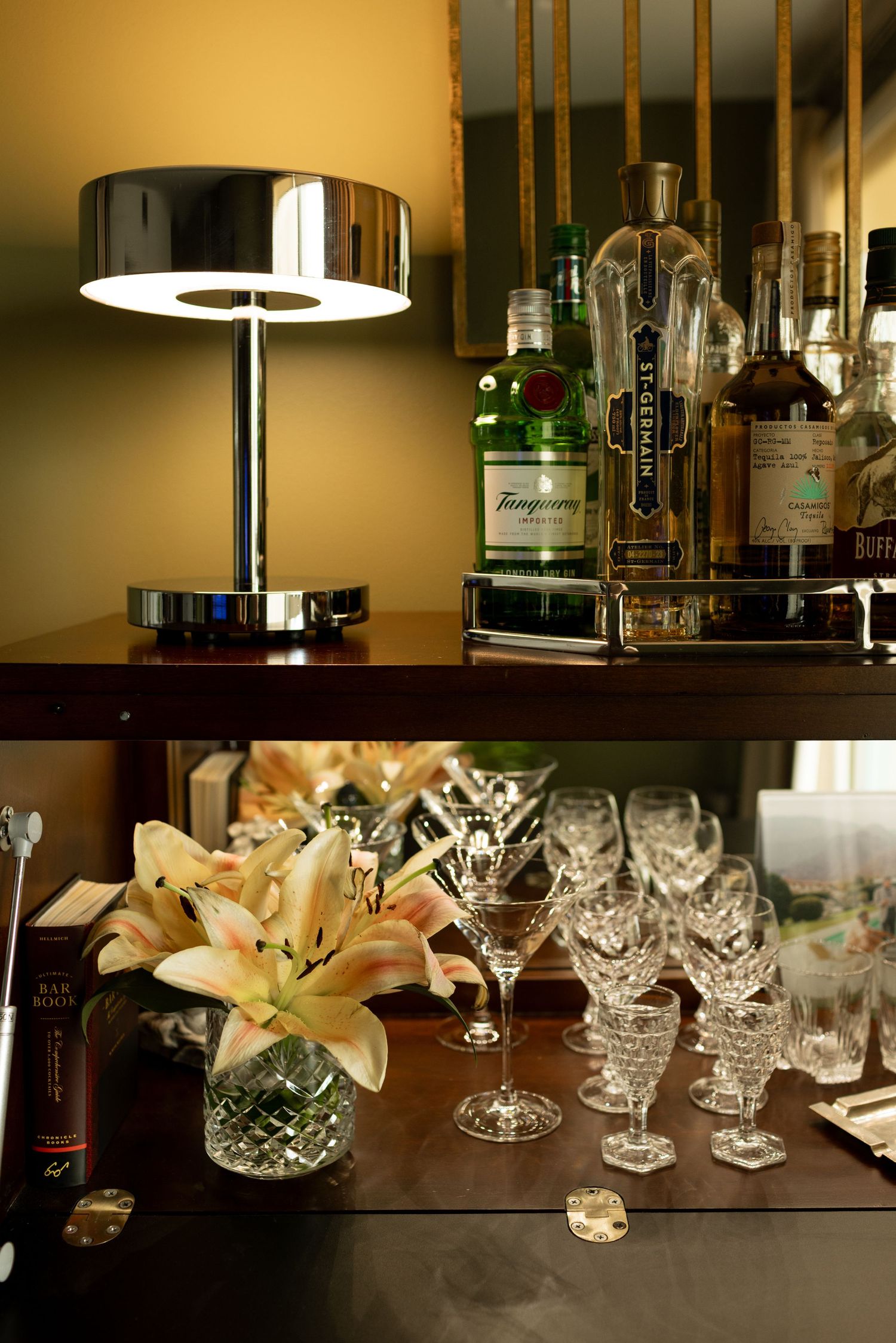 Elegant home bar with crystal glassware, fresh lilies, Tanqueray gin bottles, and a chrome lamp on dark wood shelves.