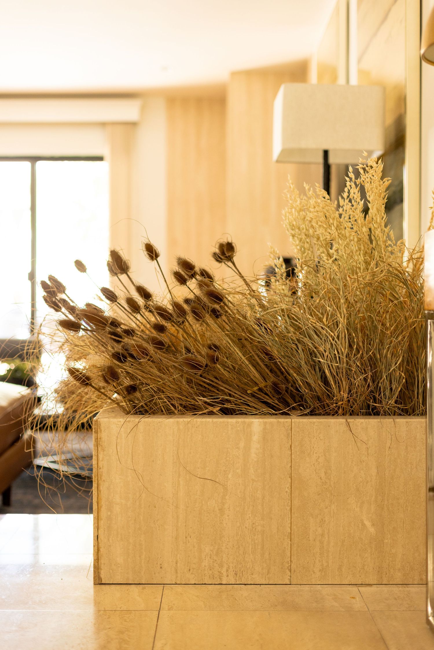 Dried ornamental grasses and thistles in a travertine marble planter box in a warm, modern interior.