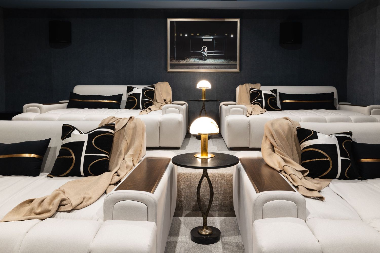 Luxury home theater with white recliner sofas, black decorative pillows, gold throw blankets, and a brass table lamp.