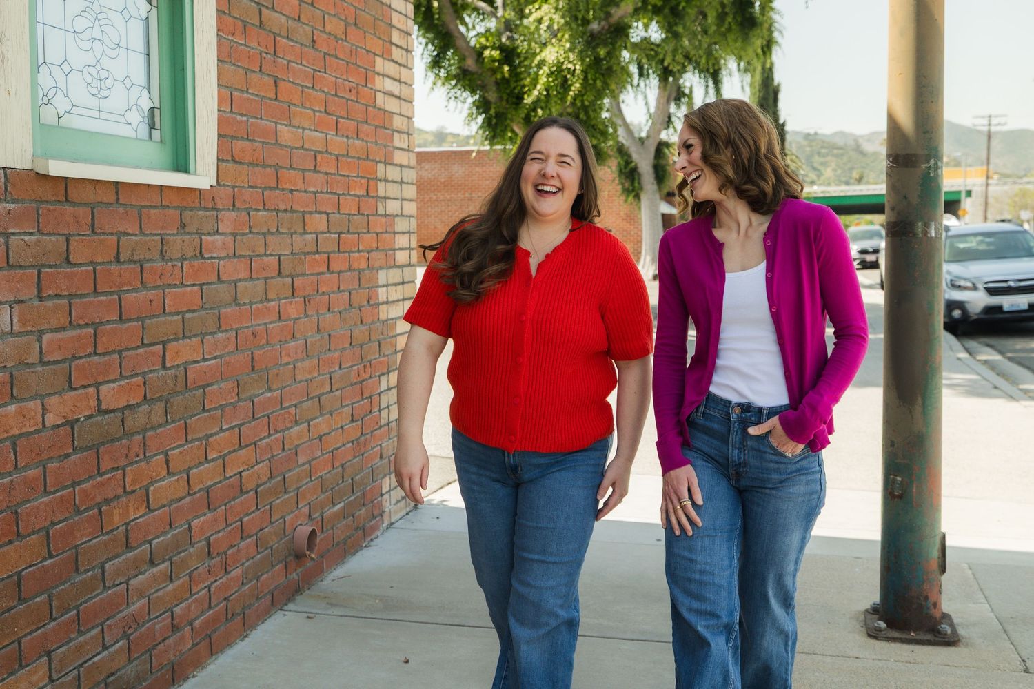 Two women in colorful tops and jeans walk and laugh together on a sunny sidewalk near a brick building.