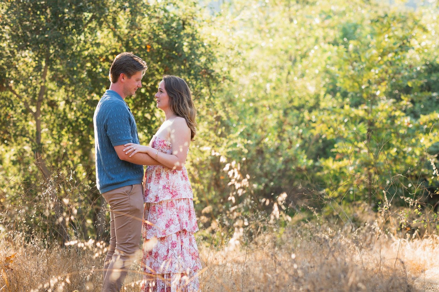 Couple embracing in a sunlit field surrounded by lush green trees, wearing casual summer attire.