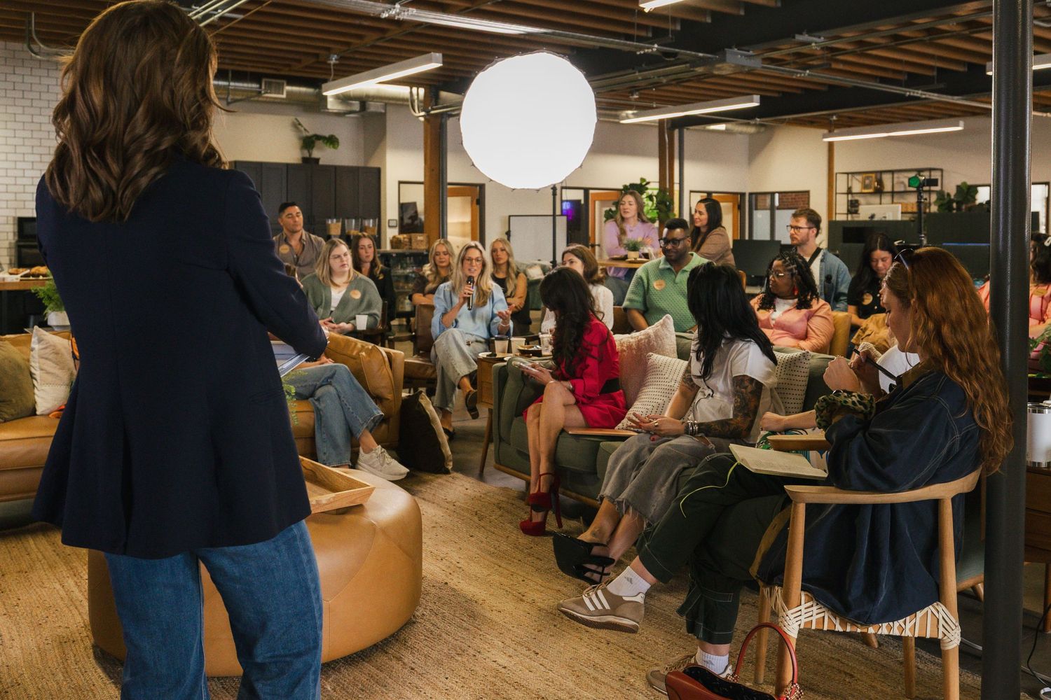 A diverse group of people attend a casual meeting in an industrial-style office space with wooden floors and exposed ceiling.