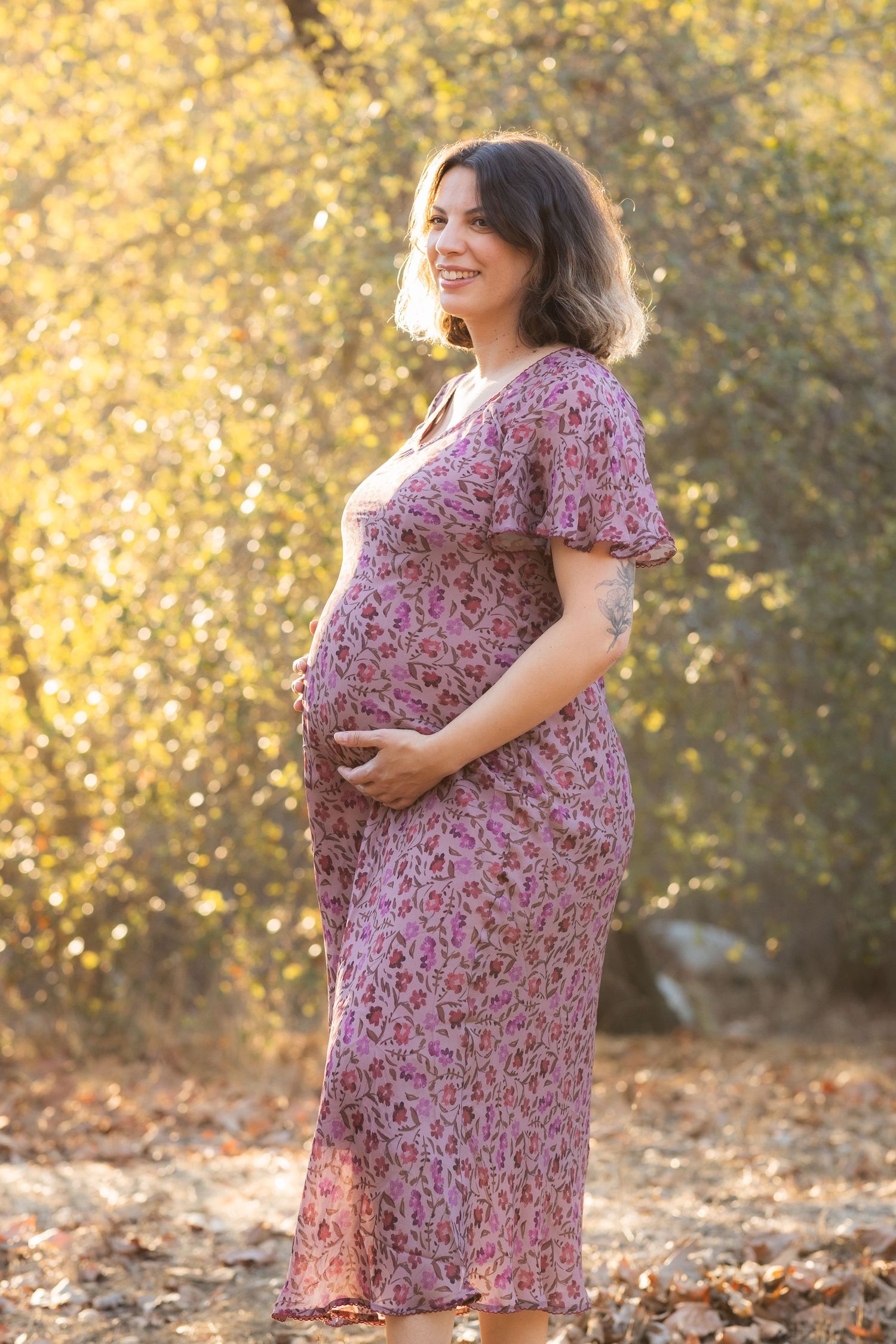 Maternity photos in floral dress against autumn foliage background.