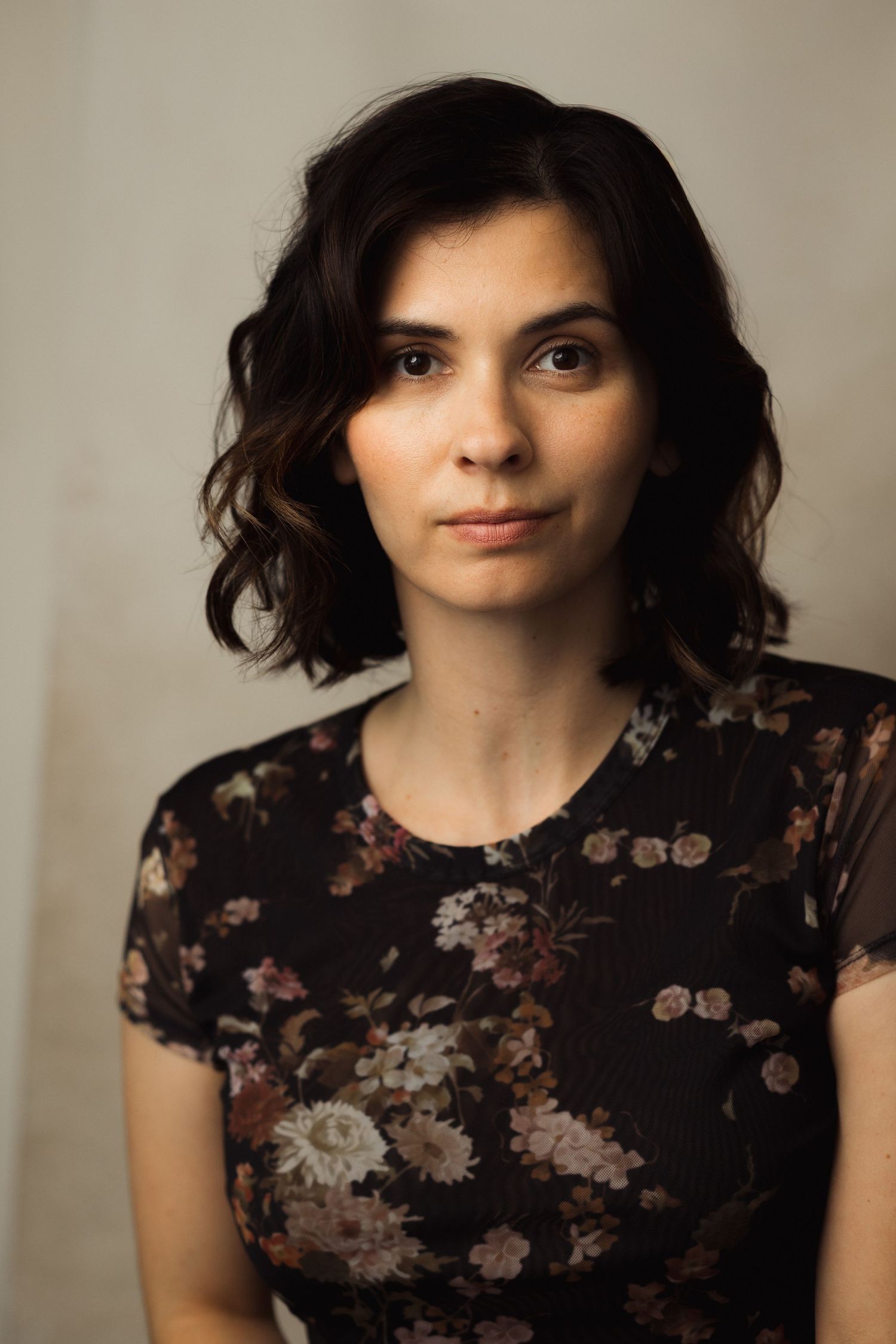 Young woman with dark wavy hair wearing a floral dress poses for a portrait against a neutral background.