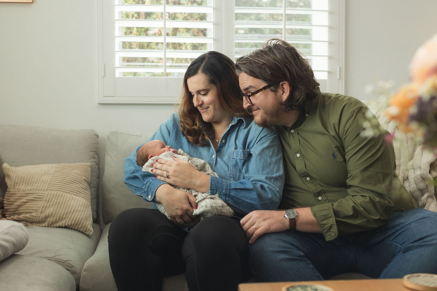 A couple sitting on a couch smiling at their newborn baby in a cozy living room setting.